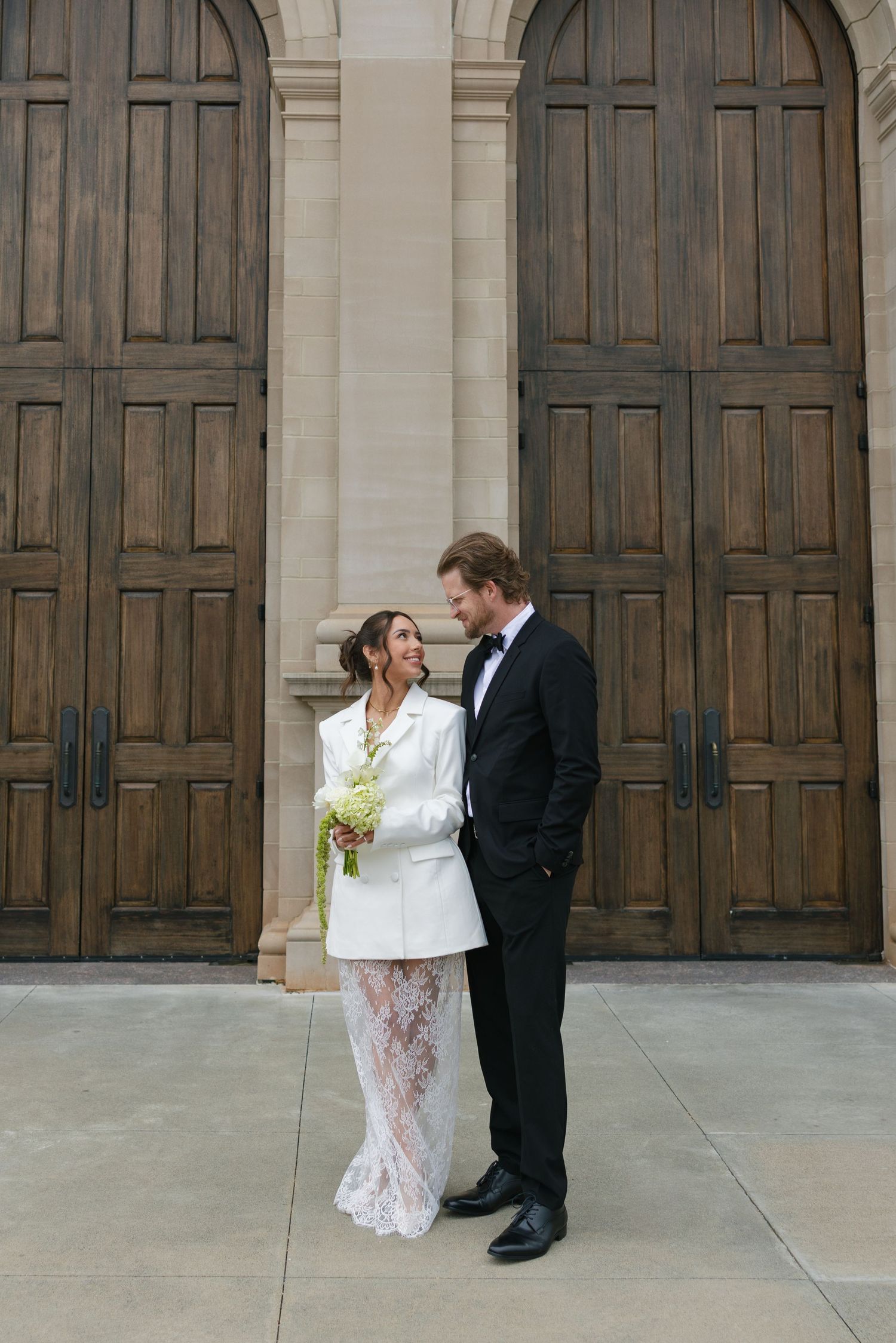 Bride in white blazer and lace skirt holding bouquet stands with groom in black tuxedo before large wooden doors.