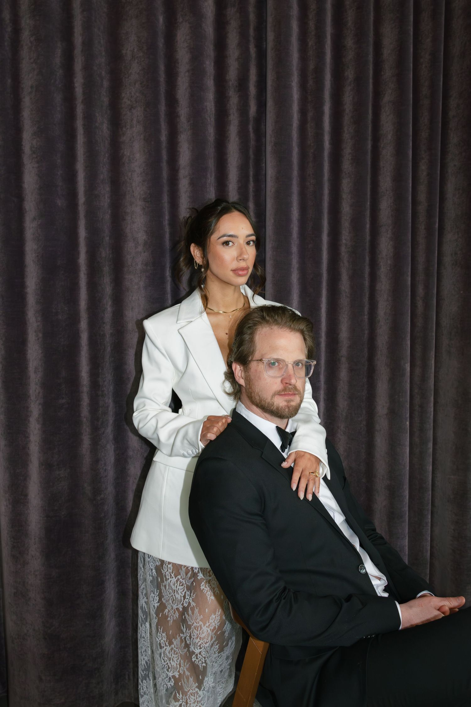 A woman in a white blazer stands behind a seated man in a black suit against a gray curtain backdrop.