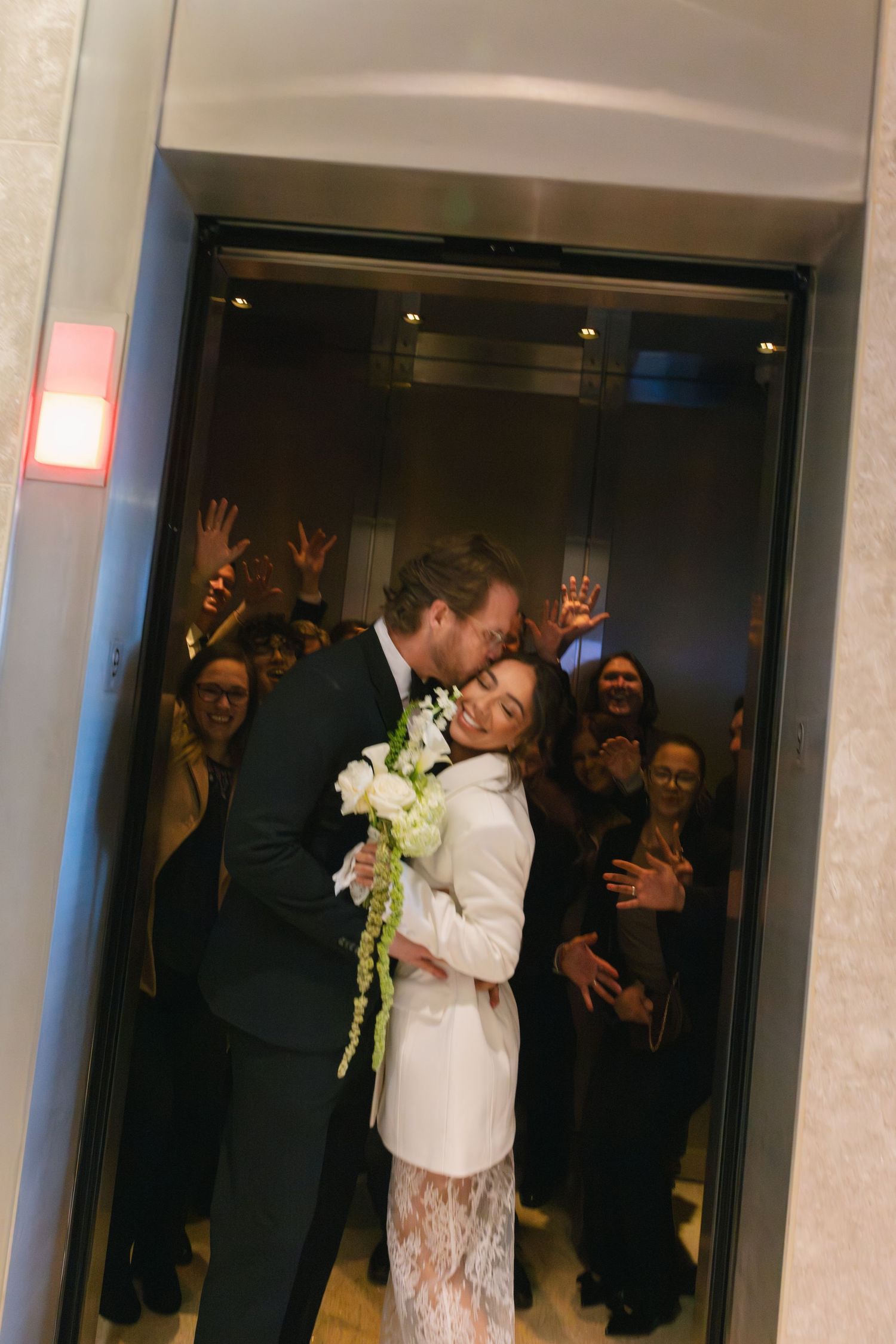 Newlywed couple sharing a kiss in elevator, bride in white lace suit holding flowers, guests celebrating behind them.