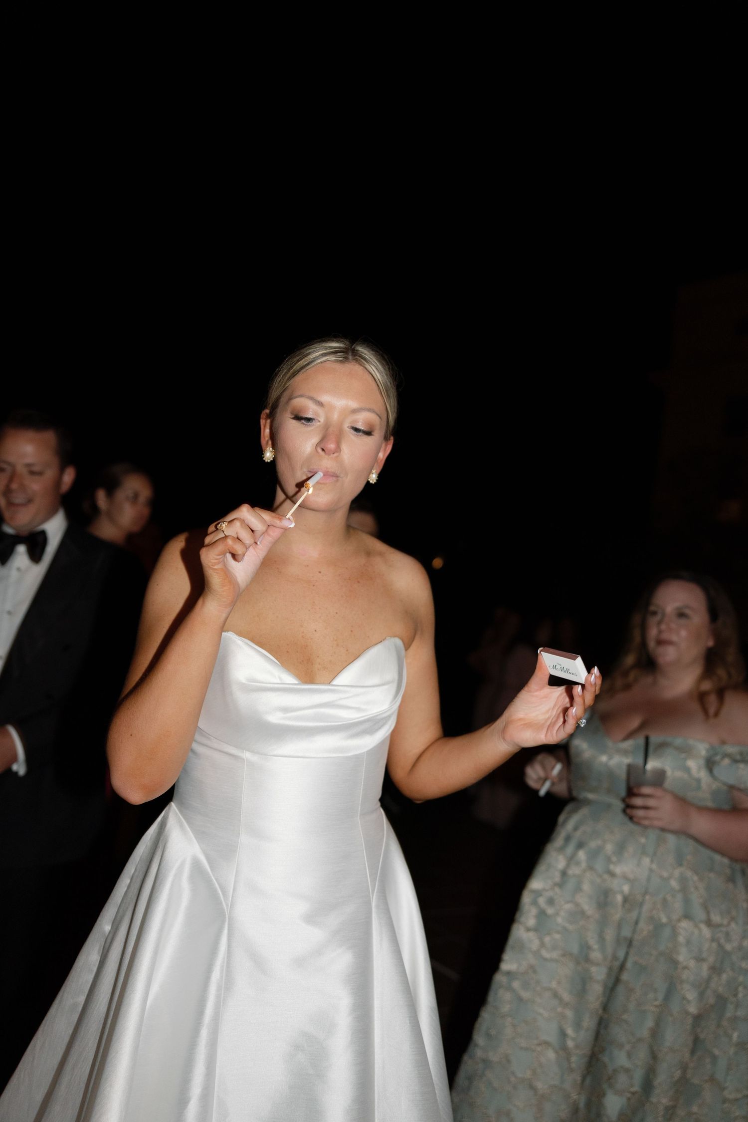Bride in white strapless gown lighting a cigarette at her wedding reception with guests in background.