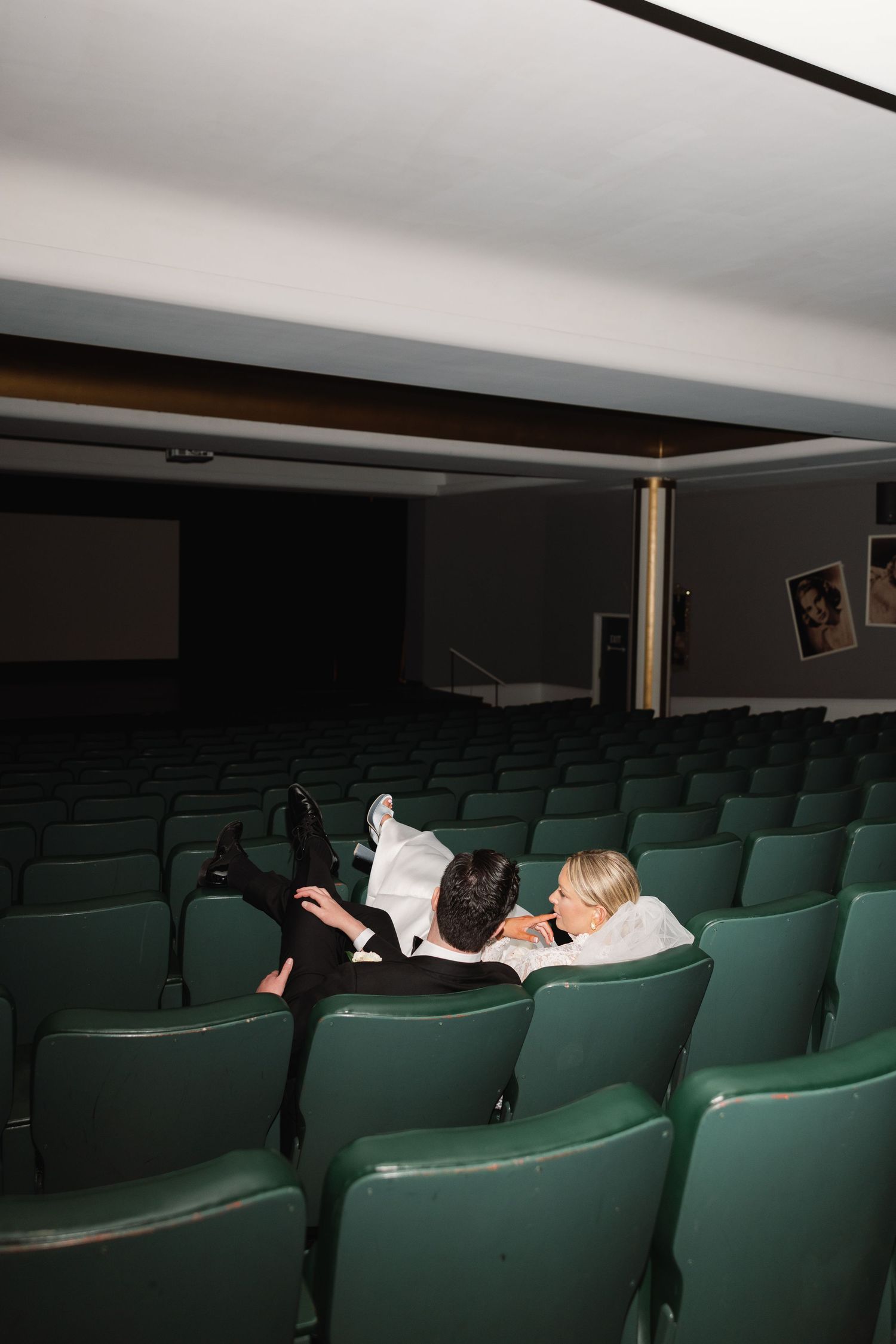 Bride and groom lying across green theater seats in an empty cinema during their wedding photo session.