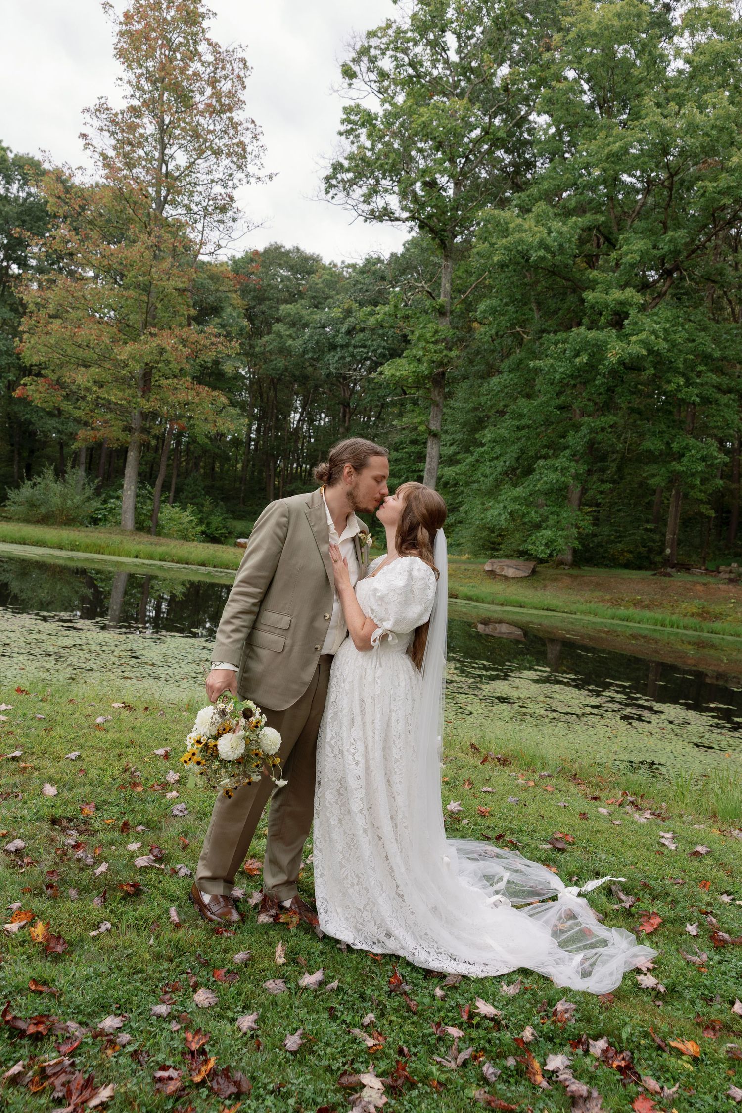 A couple shares romantic moments in a scenic garden setting with fallen autumn leaves and a pond in the background.