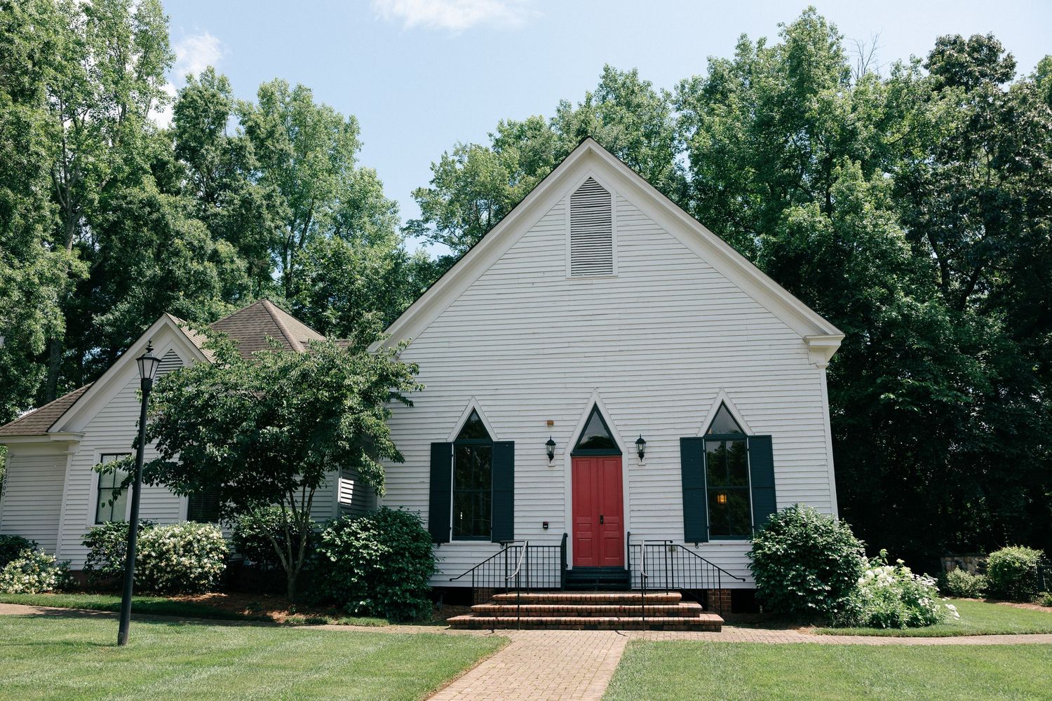 White wooden chapel with a red front door, dark green shutters, and a brick walkway, surrounded by lush green trees and landscaped shrubs.