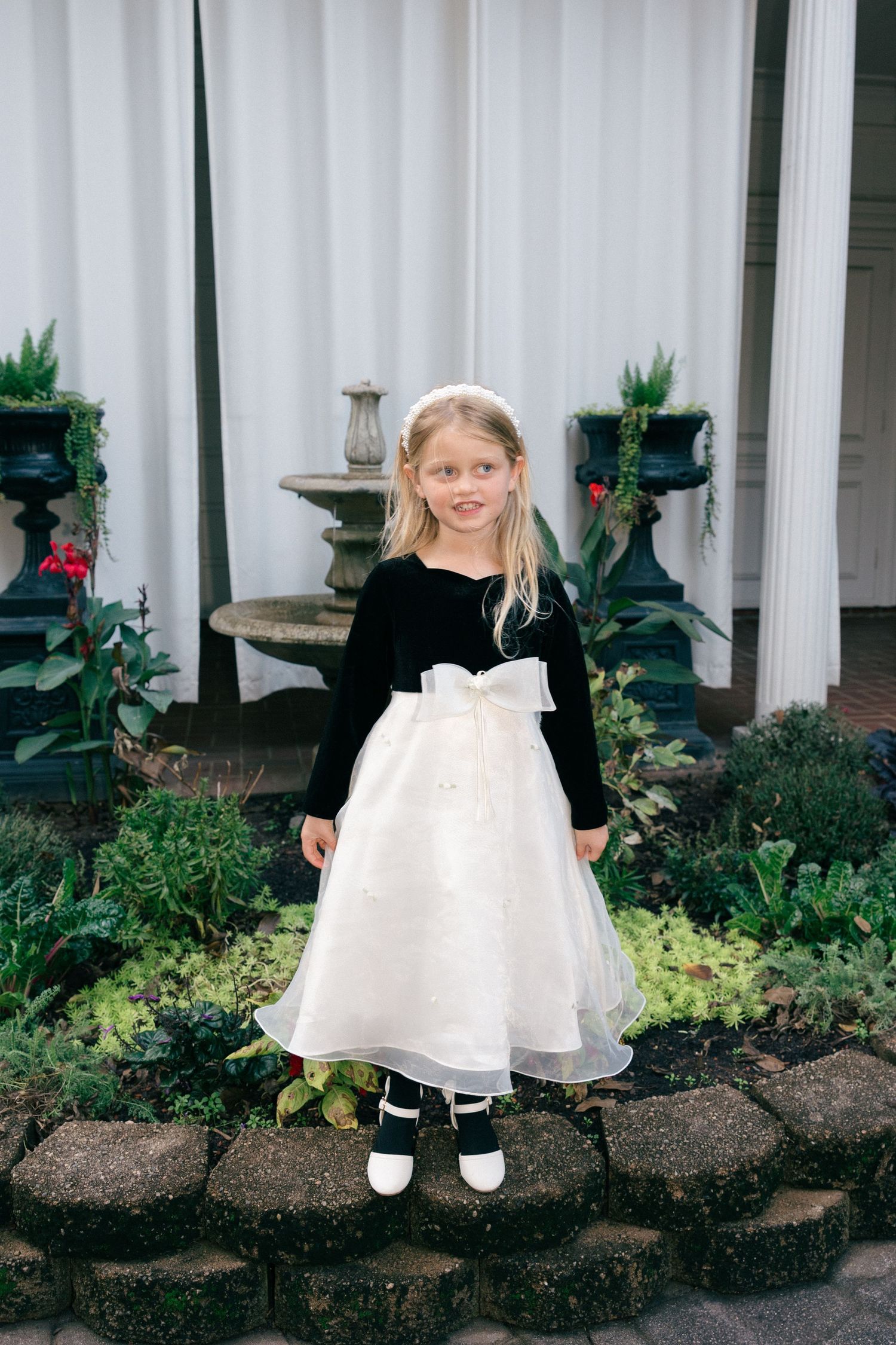 Young girl in a black velvet top and white tulle flower girl dress with a bow, standing by a garden fountain and stone border.