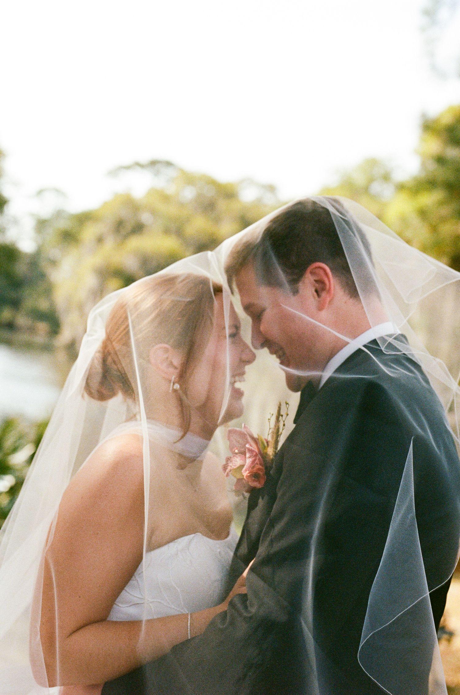 Bride and groom sharing romantic moment under wedding veil outdoors near water with lush green trees.
