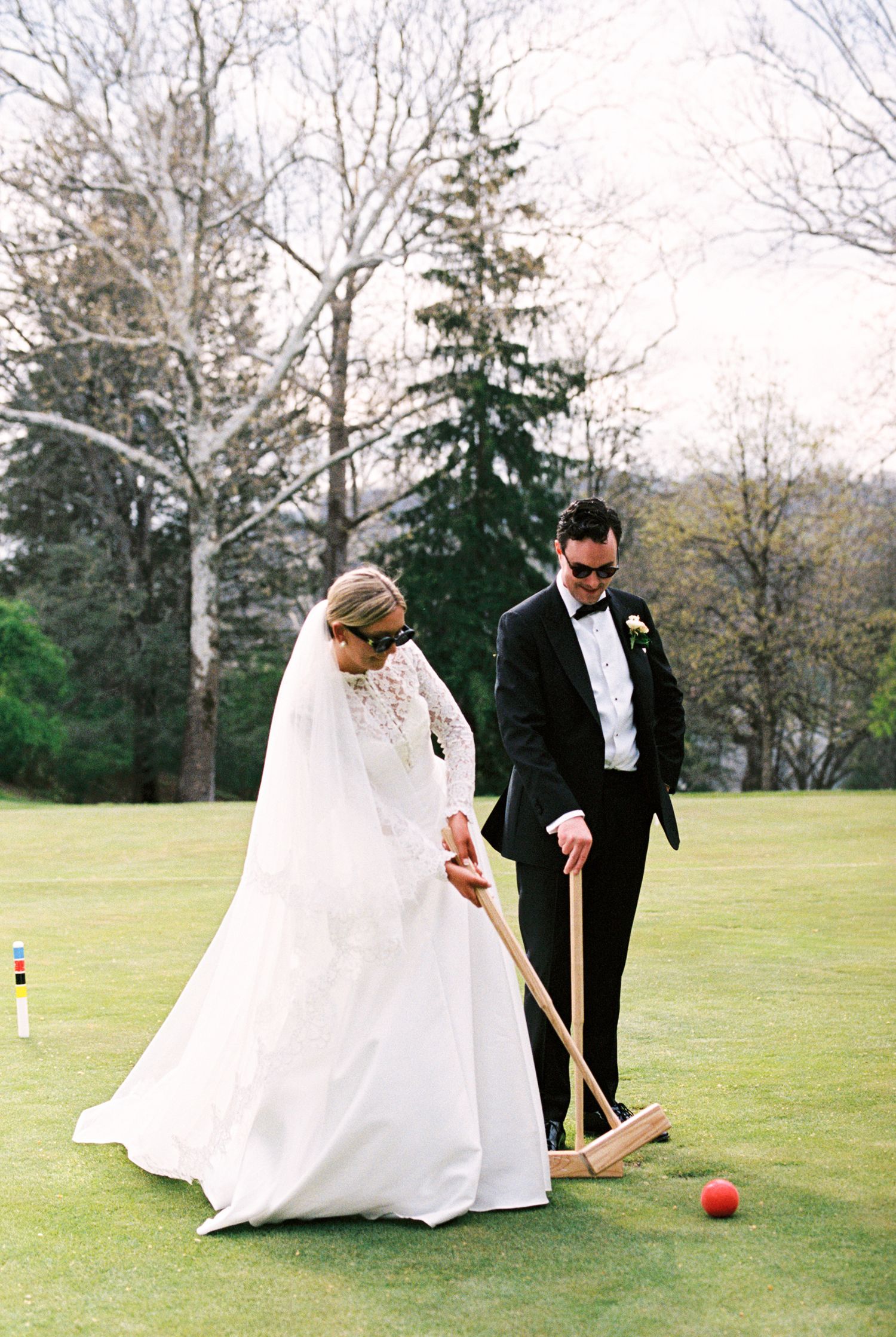 Bride and groom playing croquet on green lawn in wedding attire, white gown and black tuxedo with bow tie.