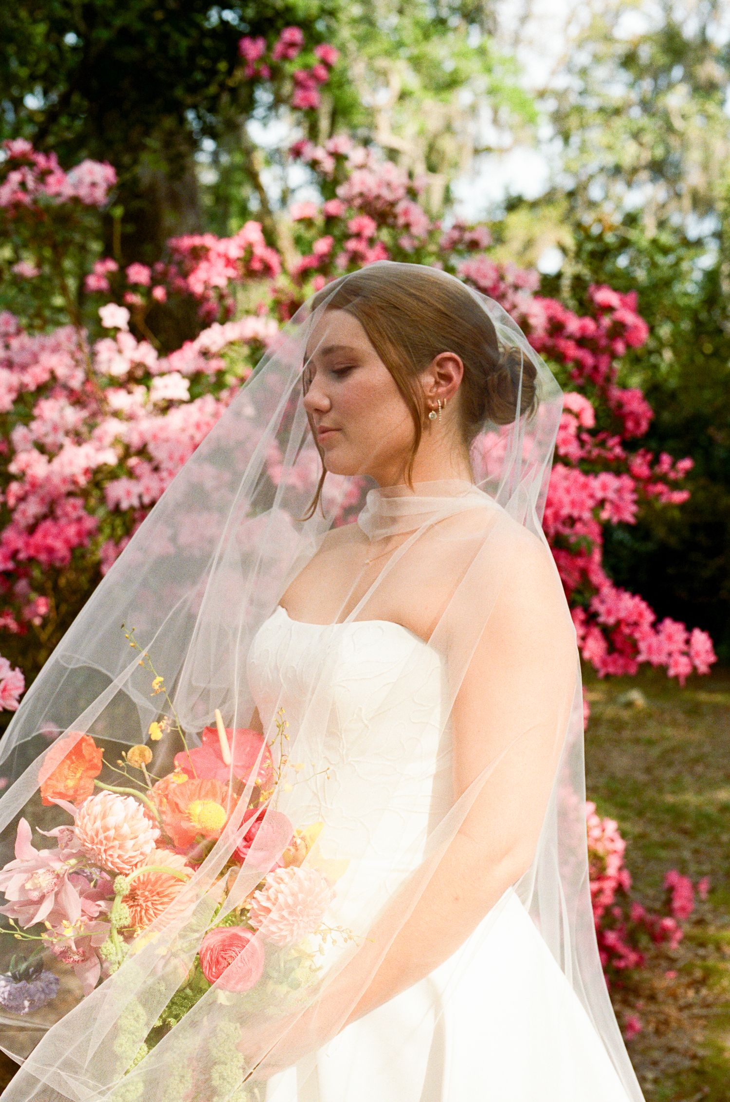Bride in white strapless gown and veil holds colorful bouquet before pink azalea blooms outdoors.