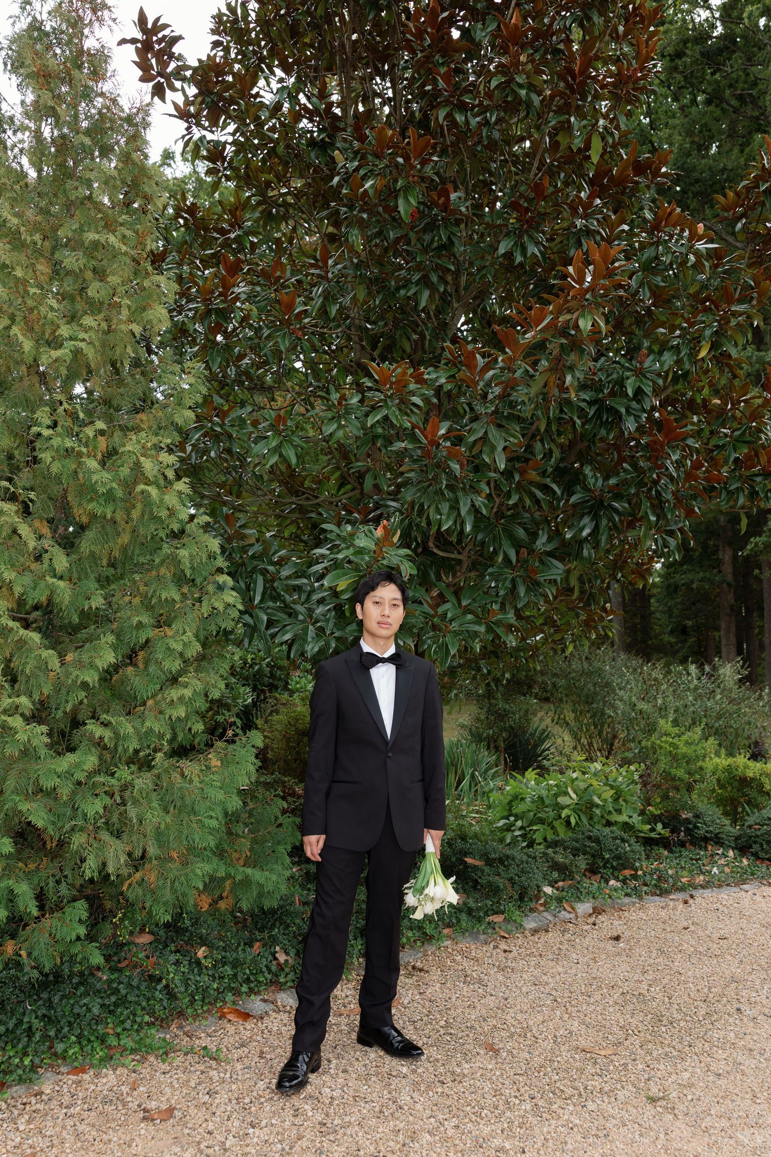 A person in formal black attire stands on a gravel path next to lush green foliage and trees in a garden setting.