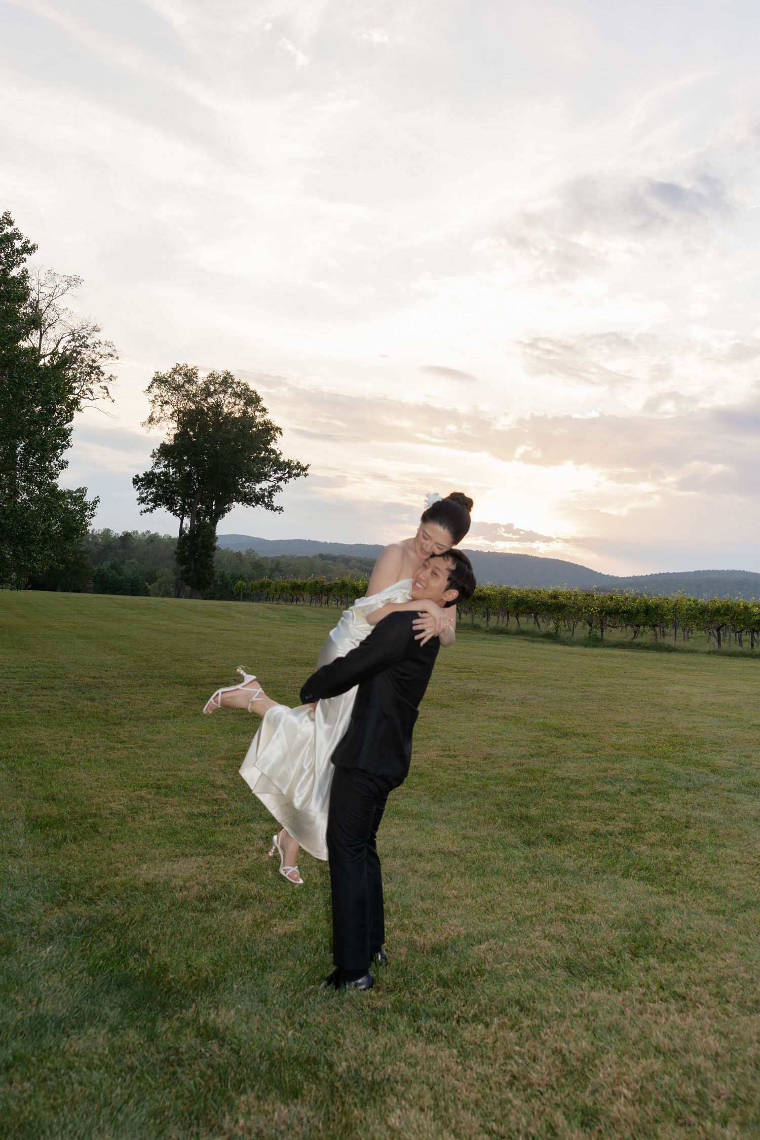 A couple shares a romantic sunset moment in formal attire on a hilltop lawn with mountains in the background.