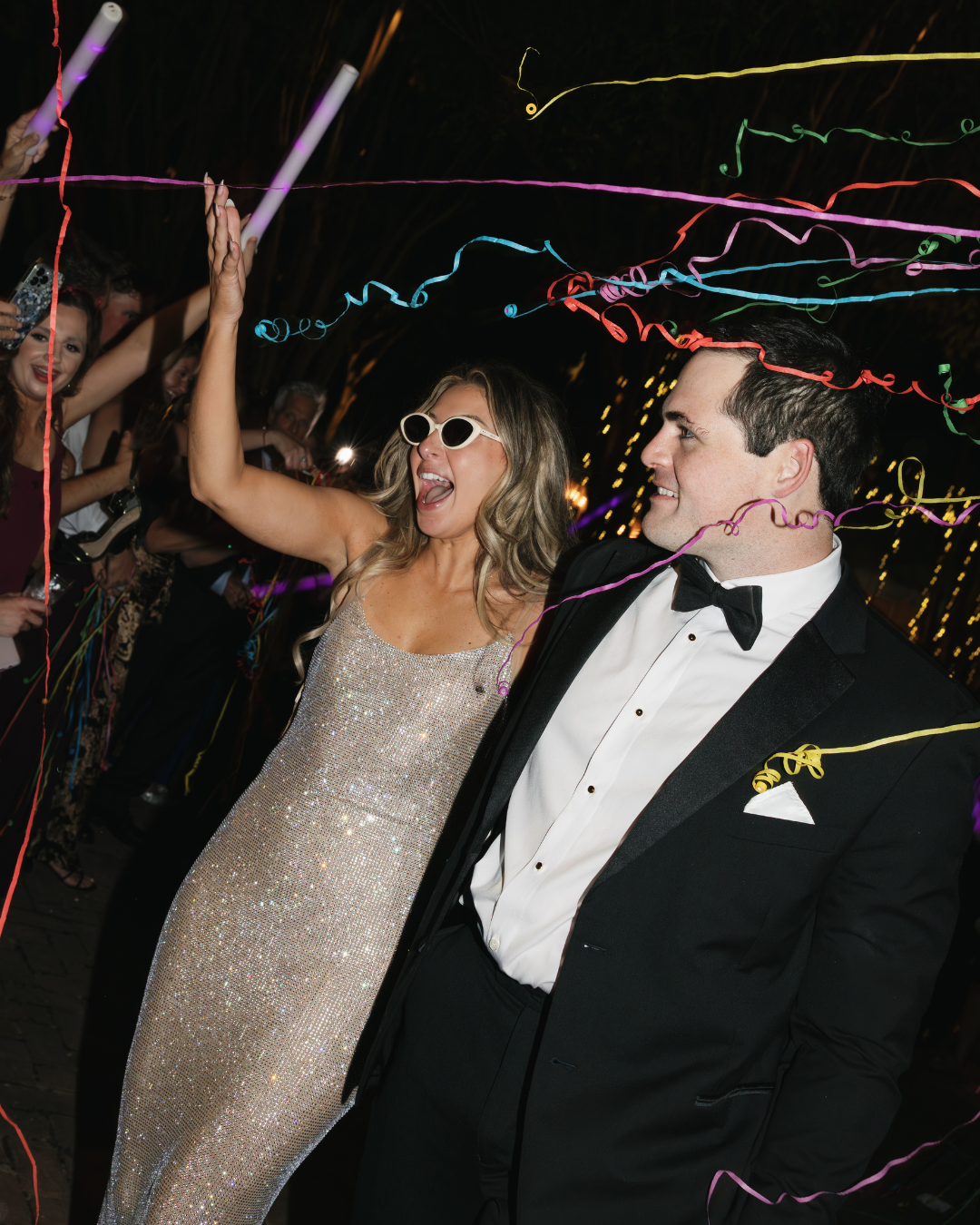 A joyful couple in wedding attire celebrates with colorful streamers and glow sticks at a festive evening reception.