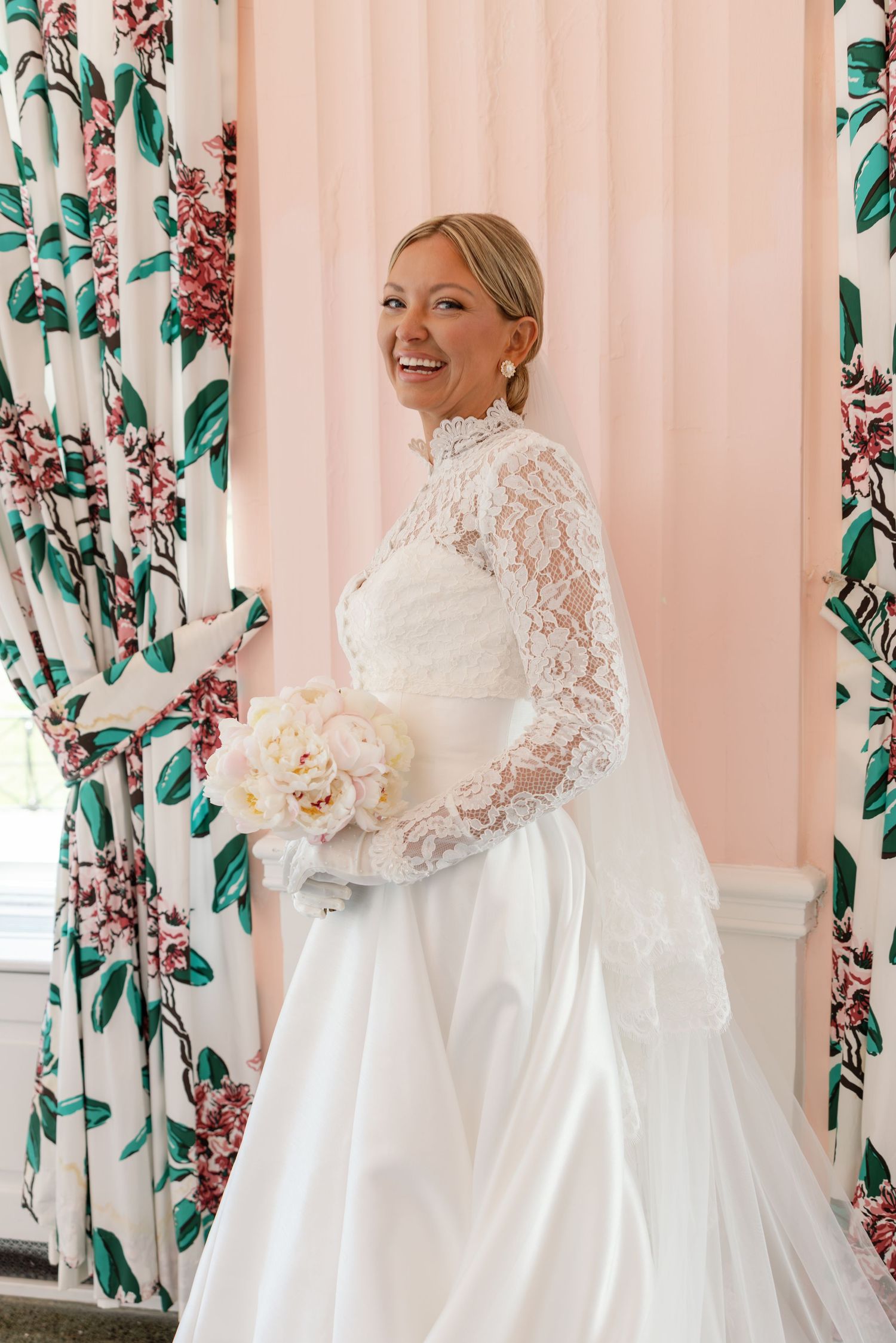 Smiling bride in lace long-sleeve wedding gown holding white peony bouquet, standing by pink wall with floral curtains.