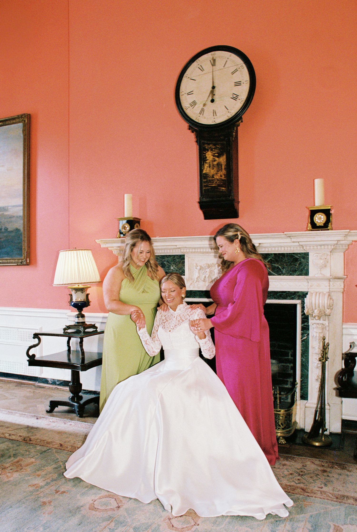 Bride seated in white gown with two bridesmaids in green and pink dresses beside a fireplace with a tall clock.