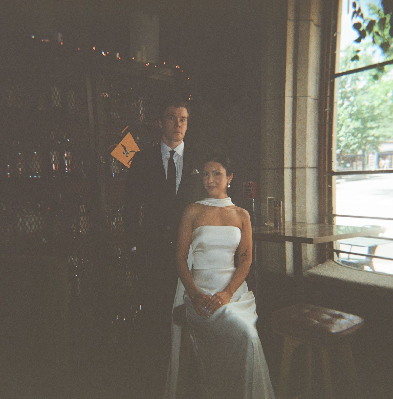Bride in a white strapless gown seated beside a groom in a dark suit and tie, posing near a sunlit window in a dimly lit vintage bar.