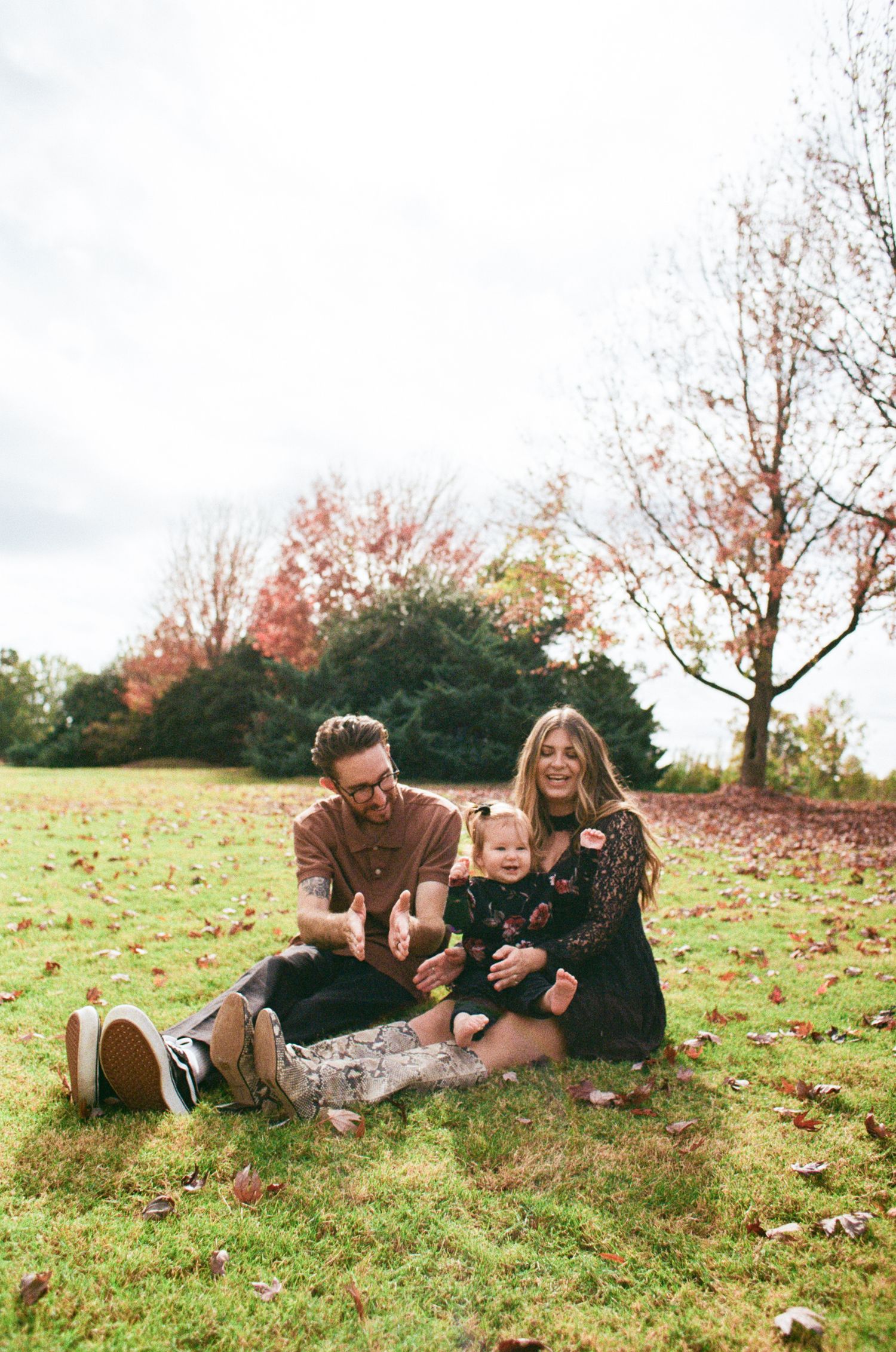Family sits together on blanket in grassy field during autumn with colorful fall trees in background.
