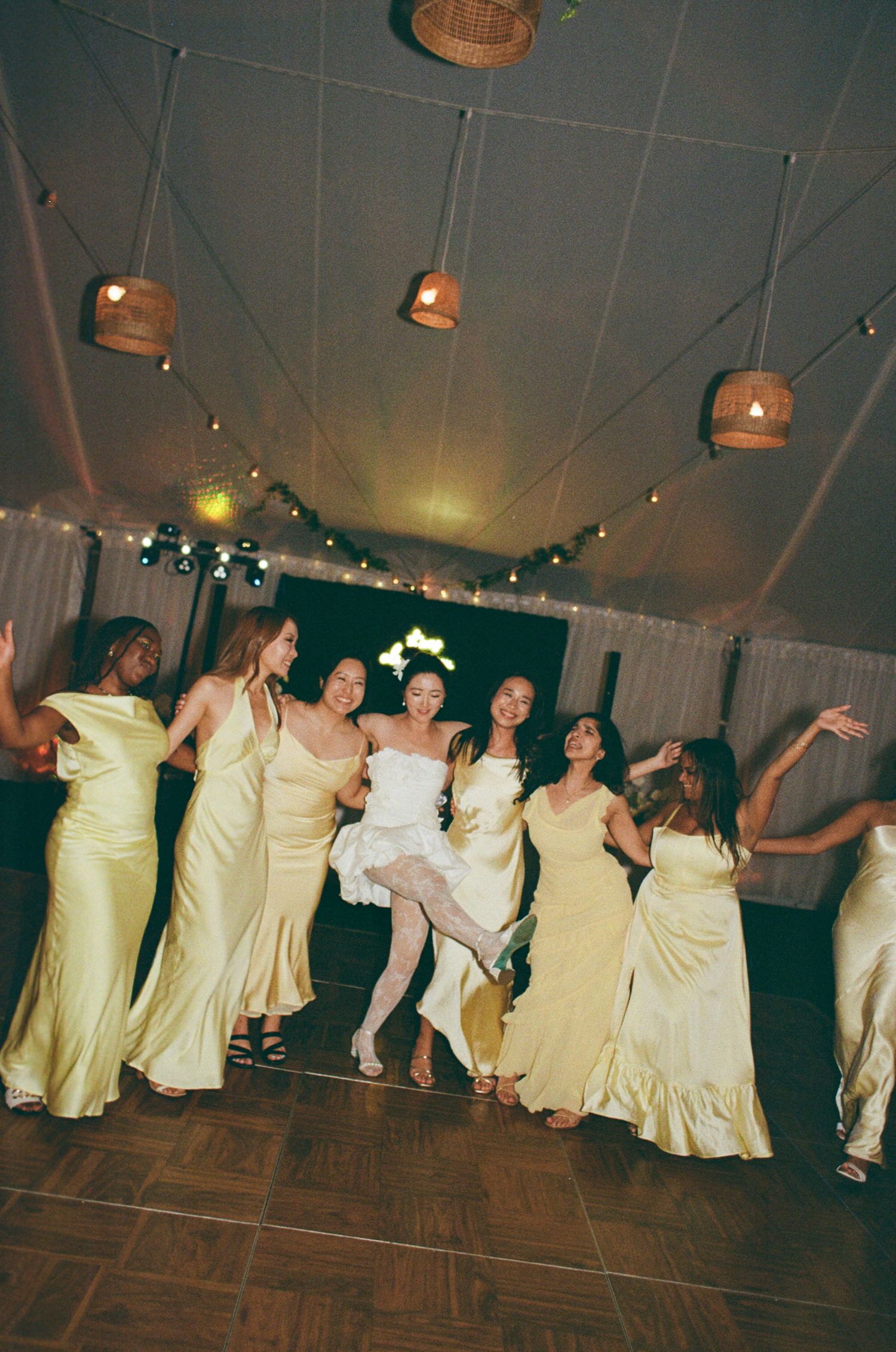 A group of people in yellow and cream colored formal dresses dance together on a wooden dance floor under hanging lights.