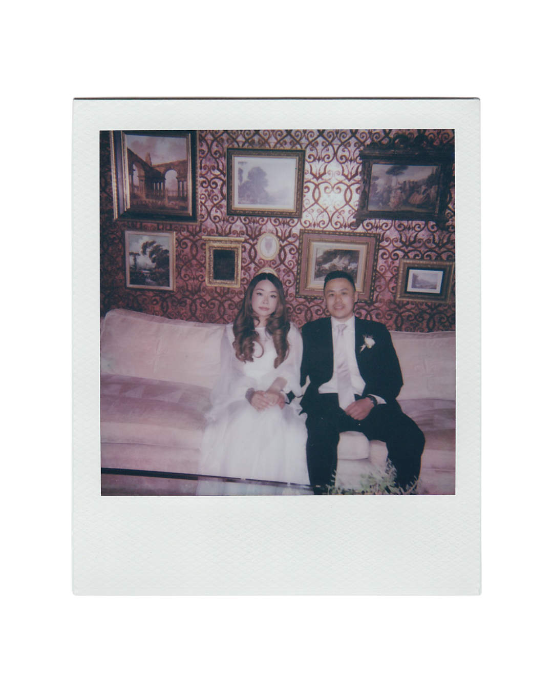 A couple in wedding attire sits together on an elegant couch in a room with ornate wallpaper and framed artwork.