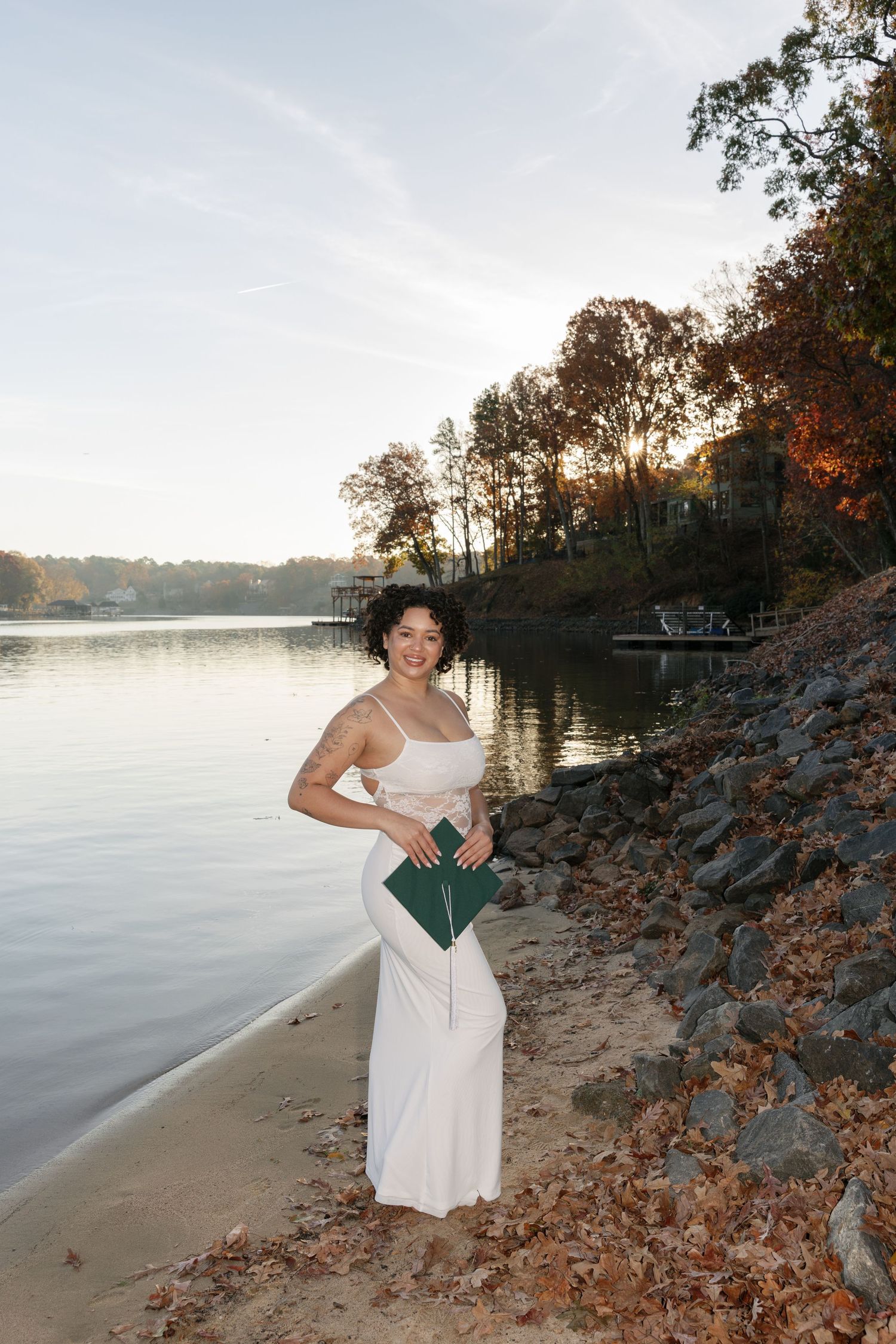 A figure in a white dress poses gracefully against a serene lakeside backdrop during autumn sunset.