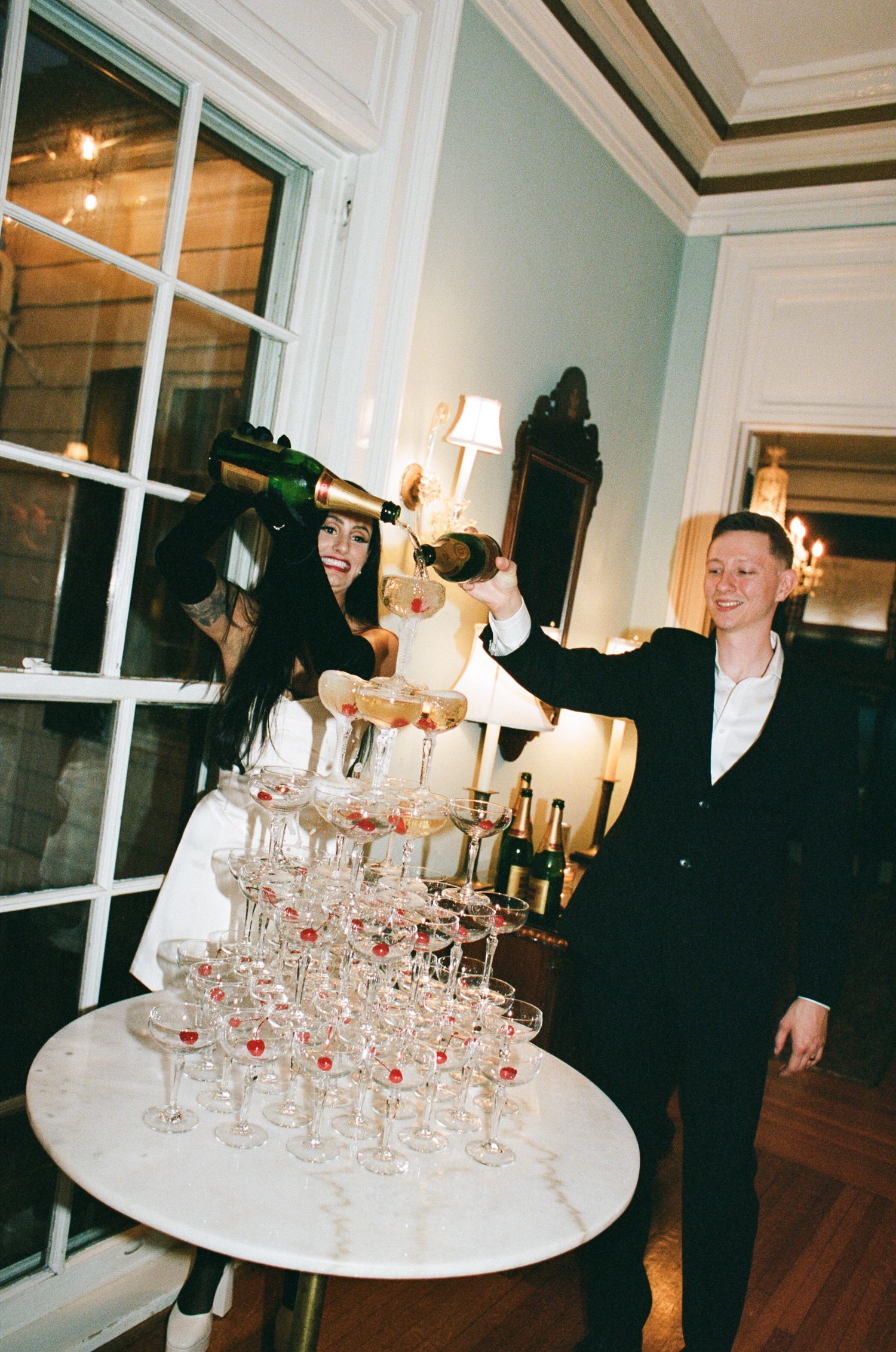Two people in formal attire pour champagne from bottles into a tall pyramid tower of coupe glasses on a marble table in an elegant room.