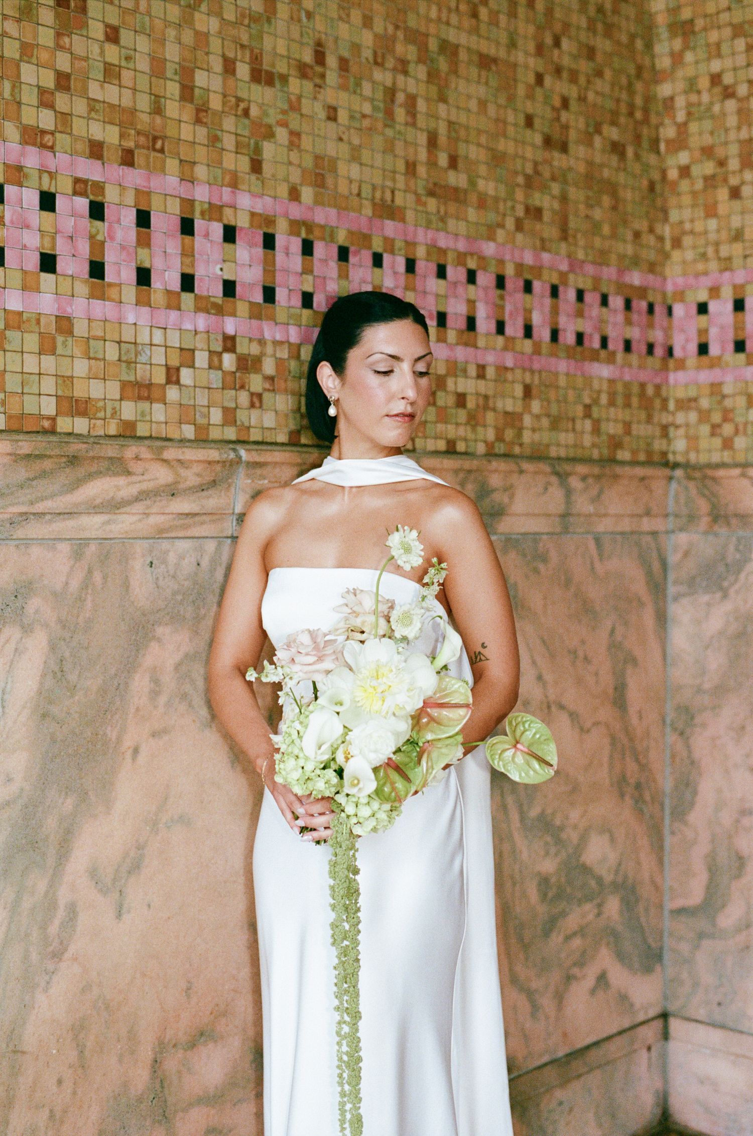 Bride in a white satin gown holding a cascading bouquet of cream blooms and anthurium against a pink marble wall with mosaic tile.