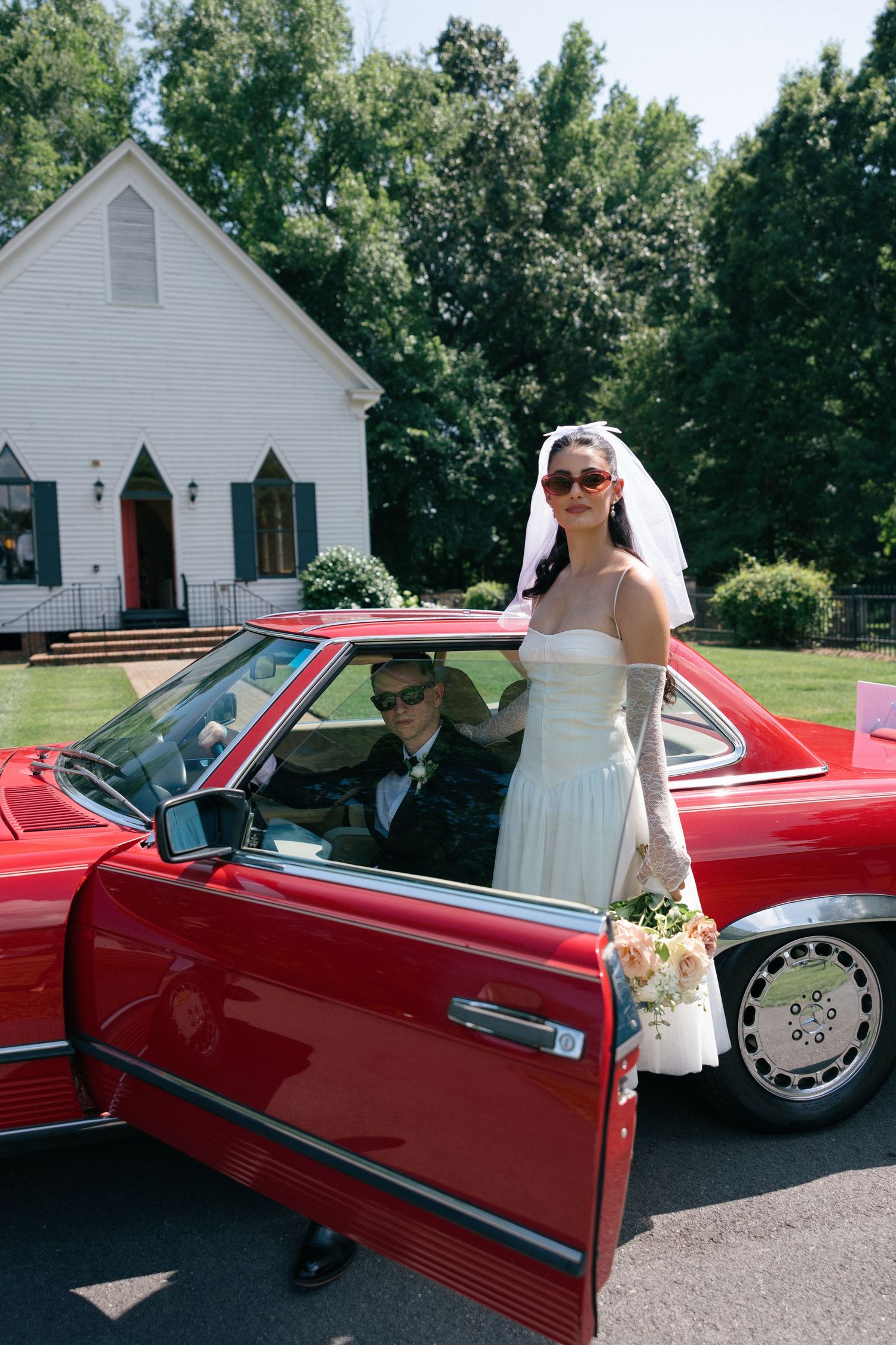 Bride in a white gown, veil and sunglasses stands beside a red vintage convertible with a groom inside, in front of a white country chapel.