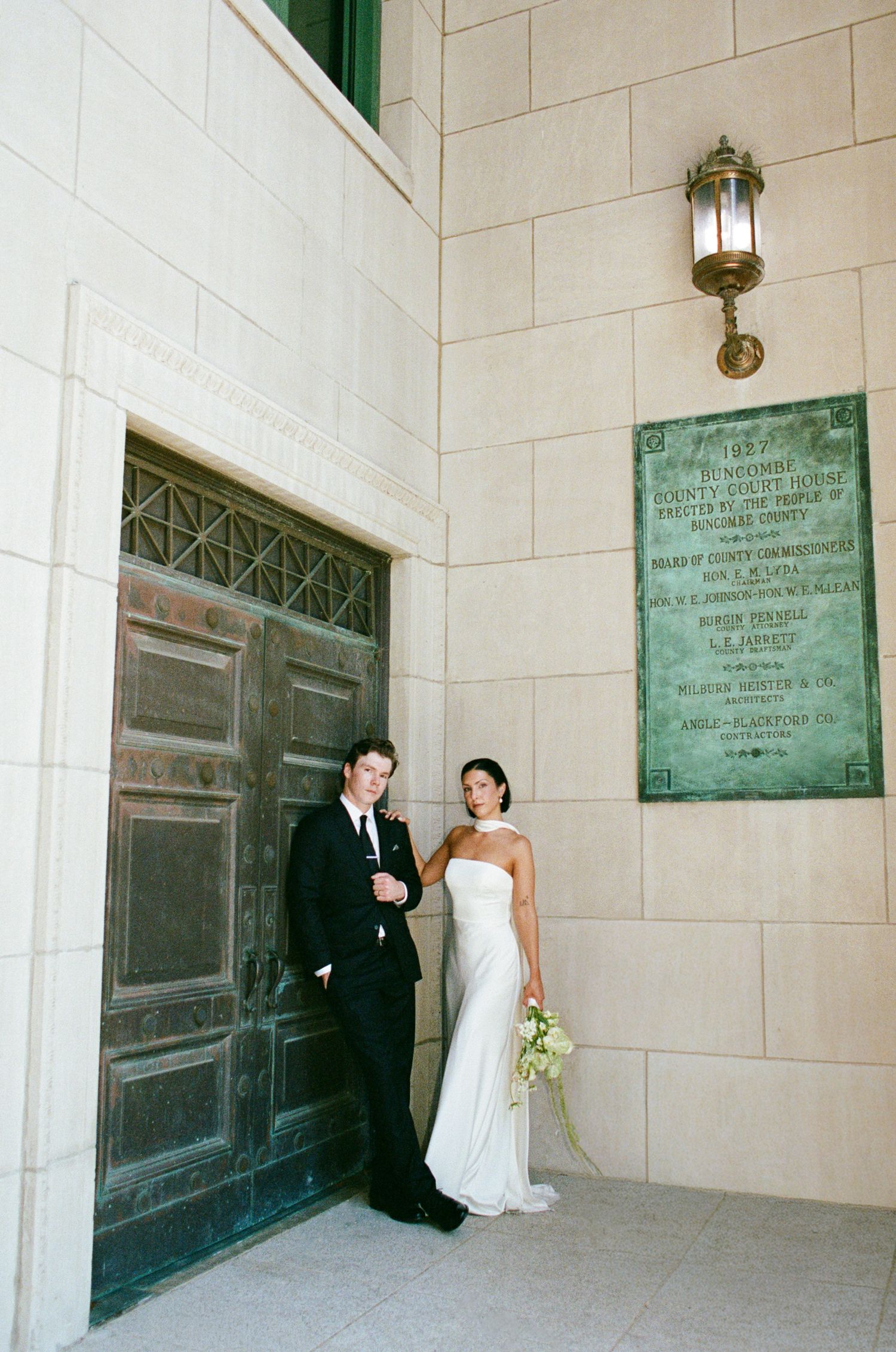 Bride in a strapless white gown holding a bouquet stands beside a groom in a black suit by an ornate bronze door and stone courthouse wall.