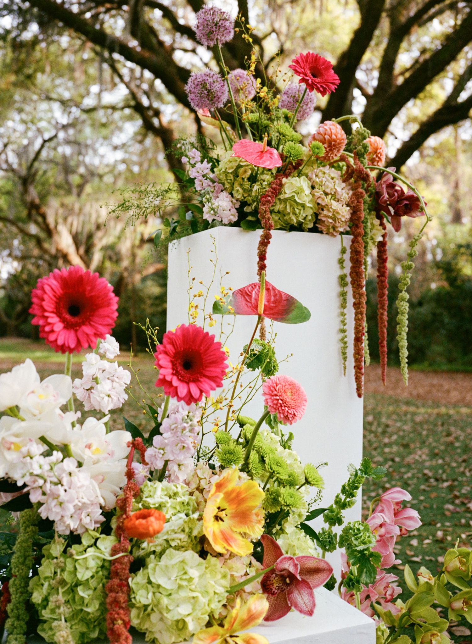 Vibrant floral arrangement with pink gerberas, hydrangeas, and tropical blooms adorning a white pedestal outdoors.