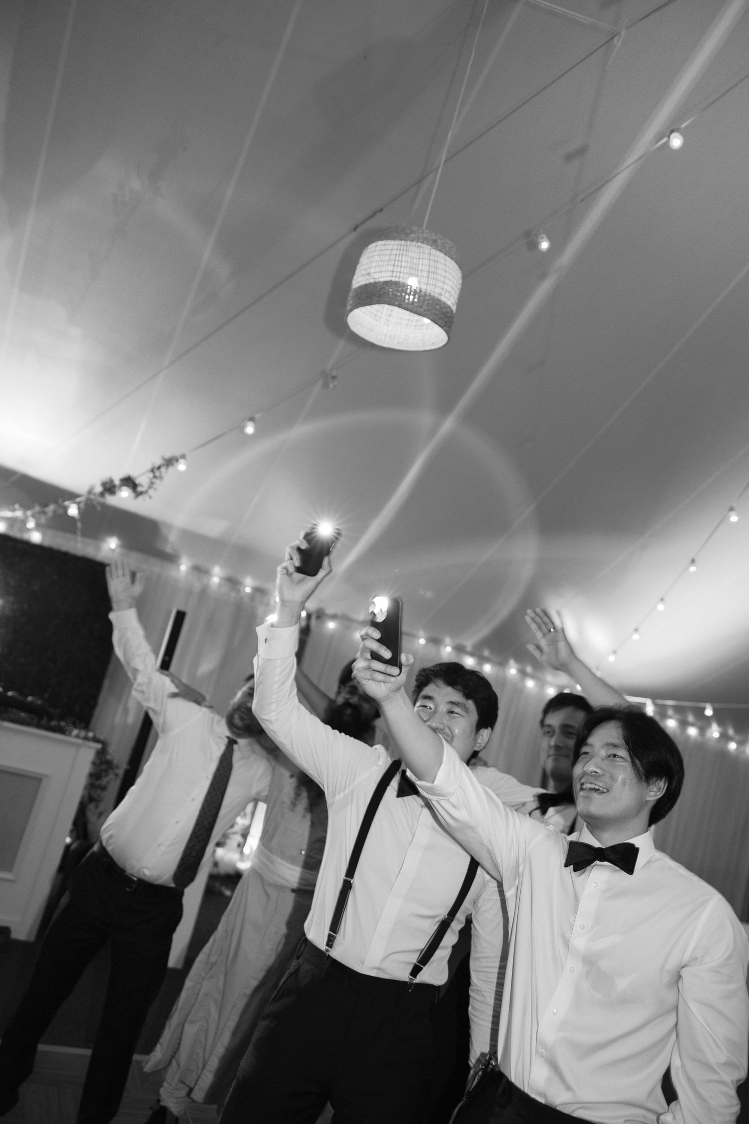 A group of people in formal attire dancing and celebrating at an indoor party with string lights overhead.