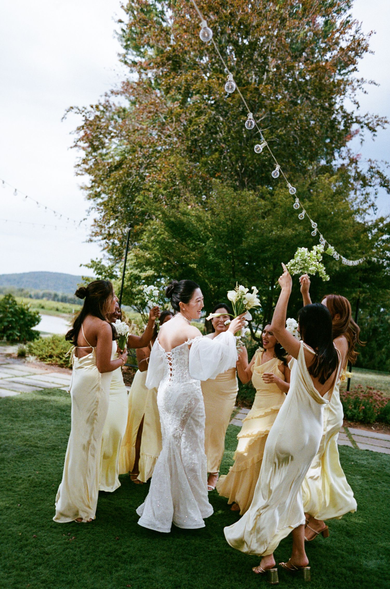 A group in flowing cream and white dresses dances joyfully on a grassy hillside during a wedding celebration.