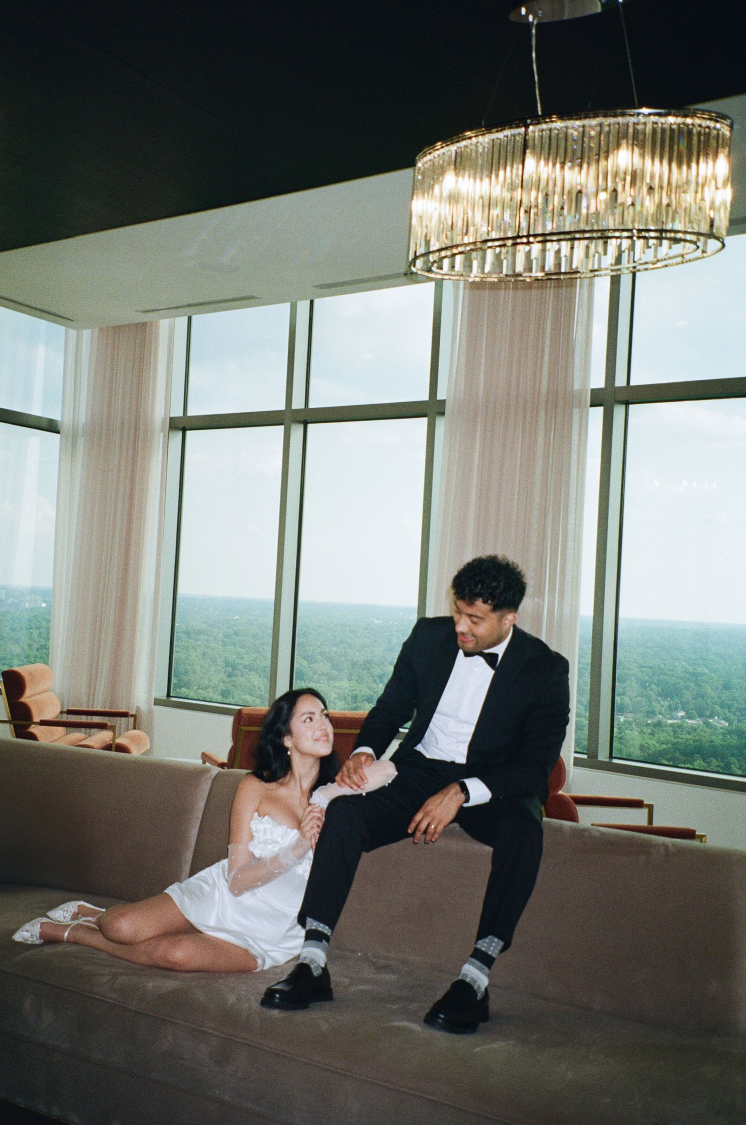 A couple in formal attire sits together in an elegant room with floor-to-ceiling windows and a crystal chandelier.