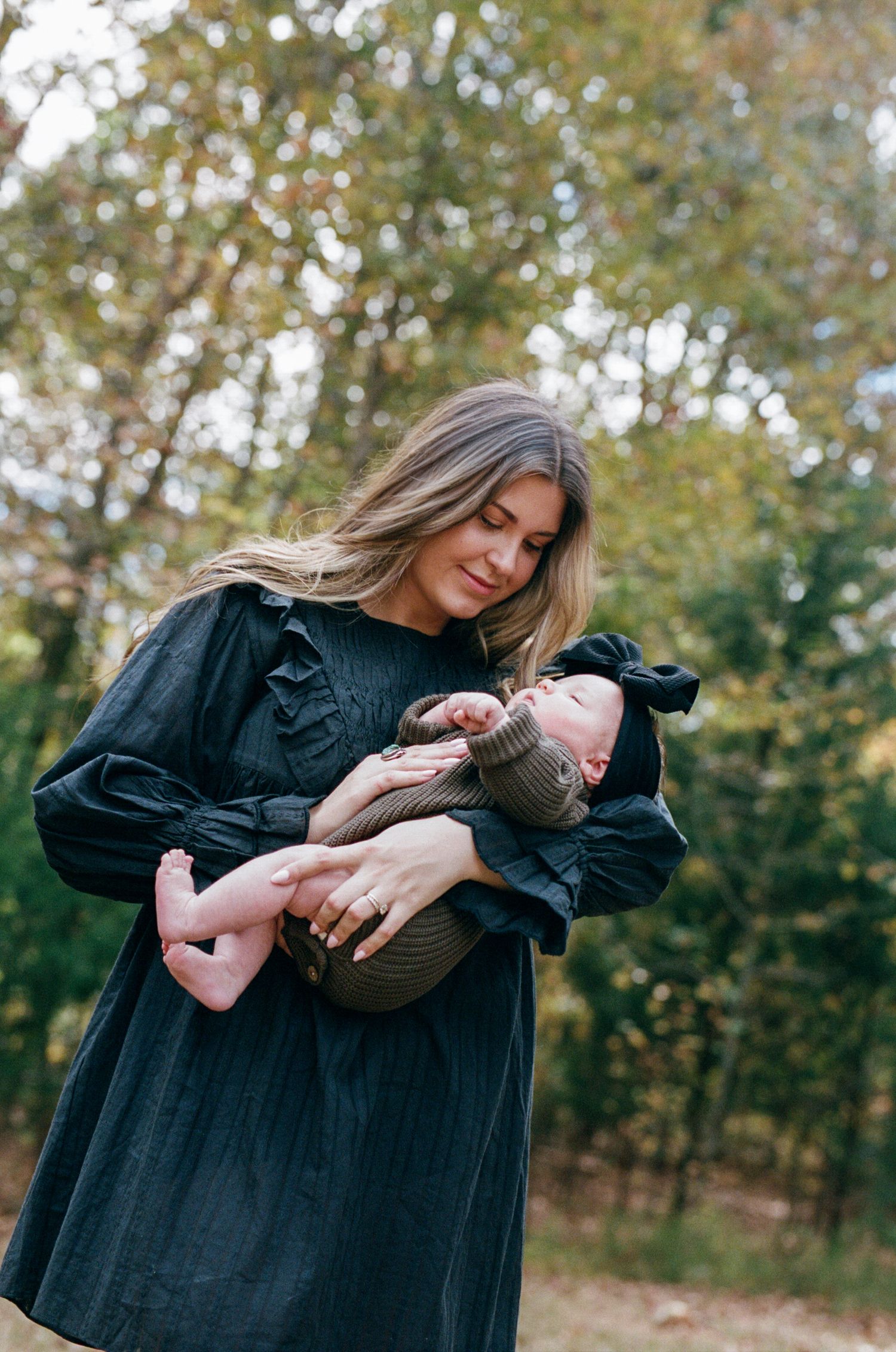 A woman in a black dress holds a baby wearing a brown sweater outdoors among autumn trees.