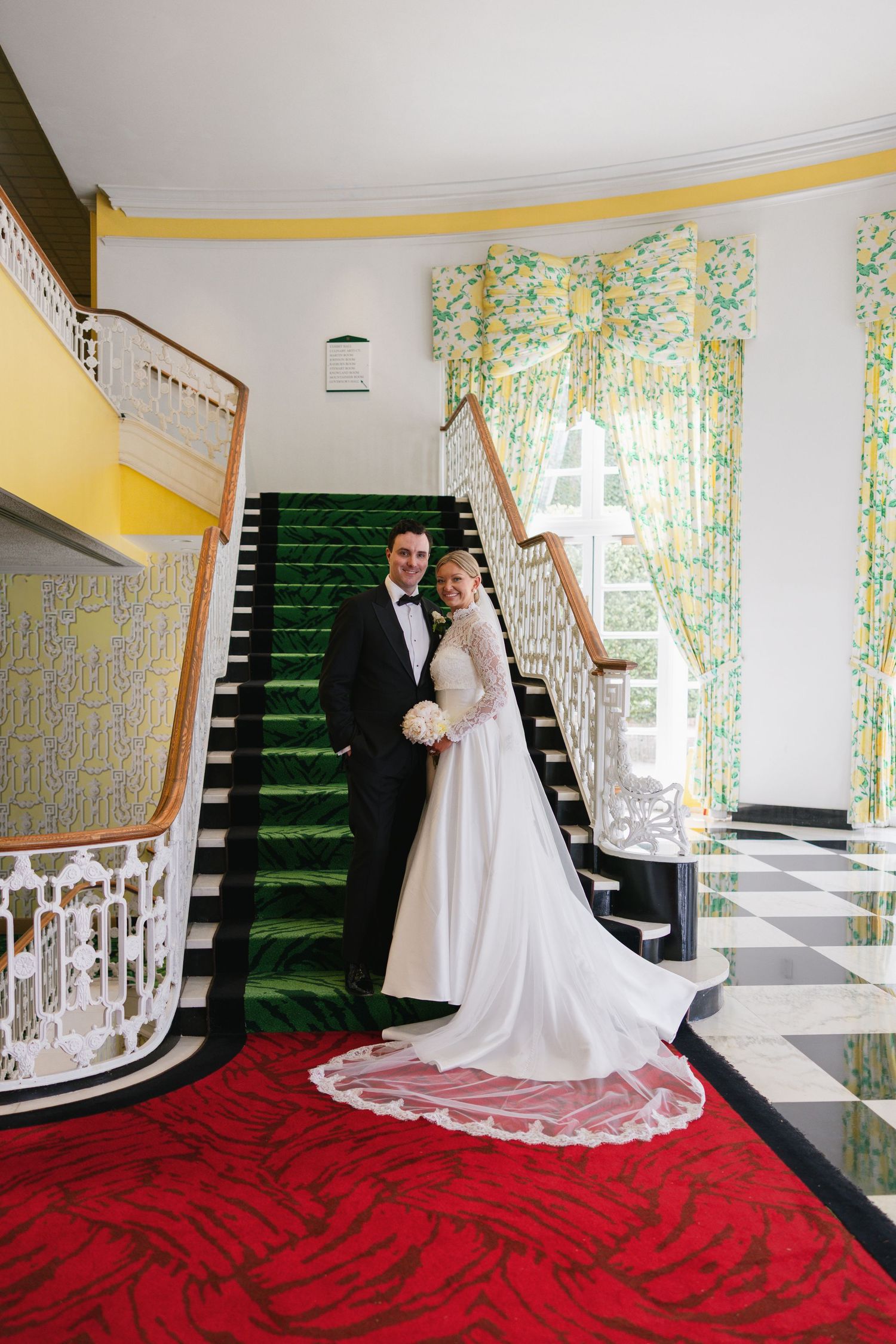 Bride and groom posing elegantly on green carpeted staircase in grand venue with checkered floors.