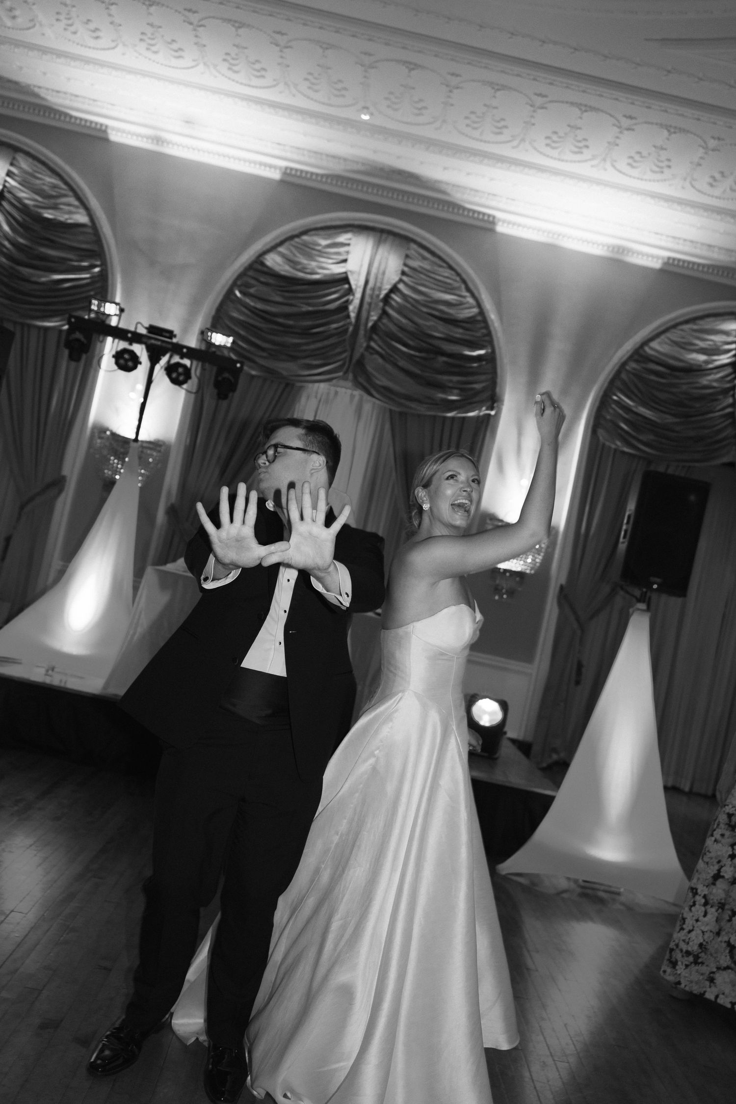 Bride and groom dancing joyfully at wedding reception in elegant ballroom, black and white photo.