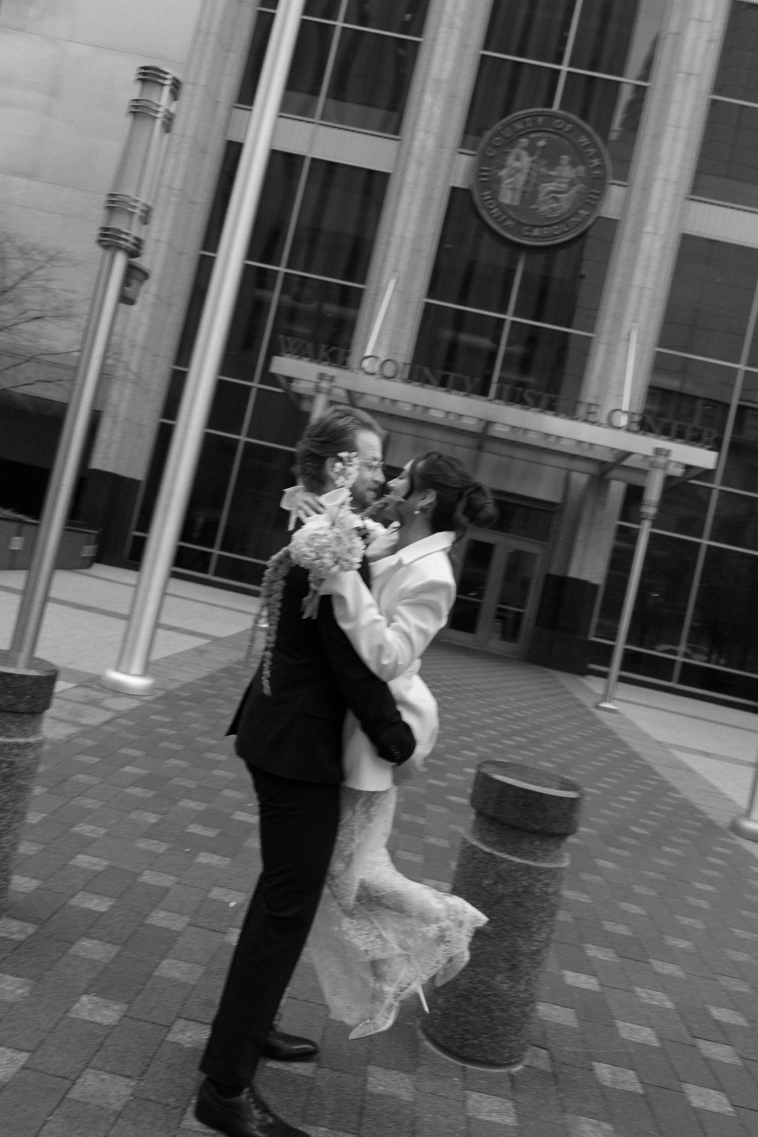 Black and white photo of a couple embracing and kissing outside a civic center building on their wedding day.