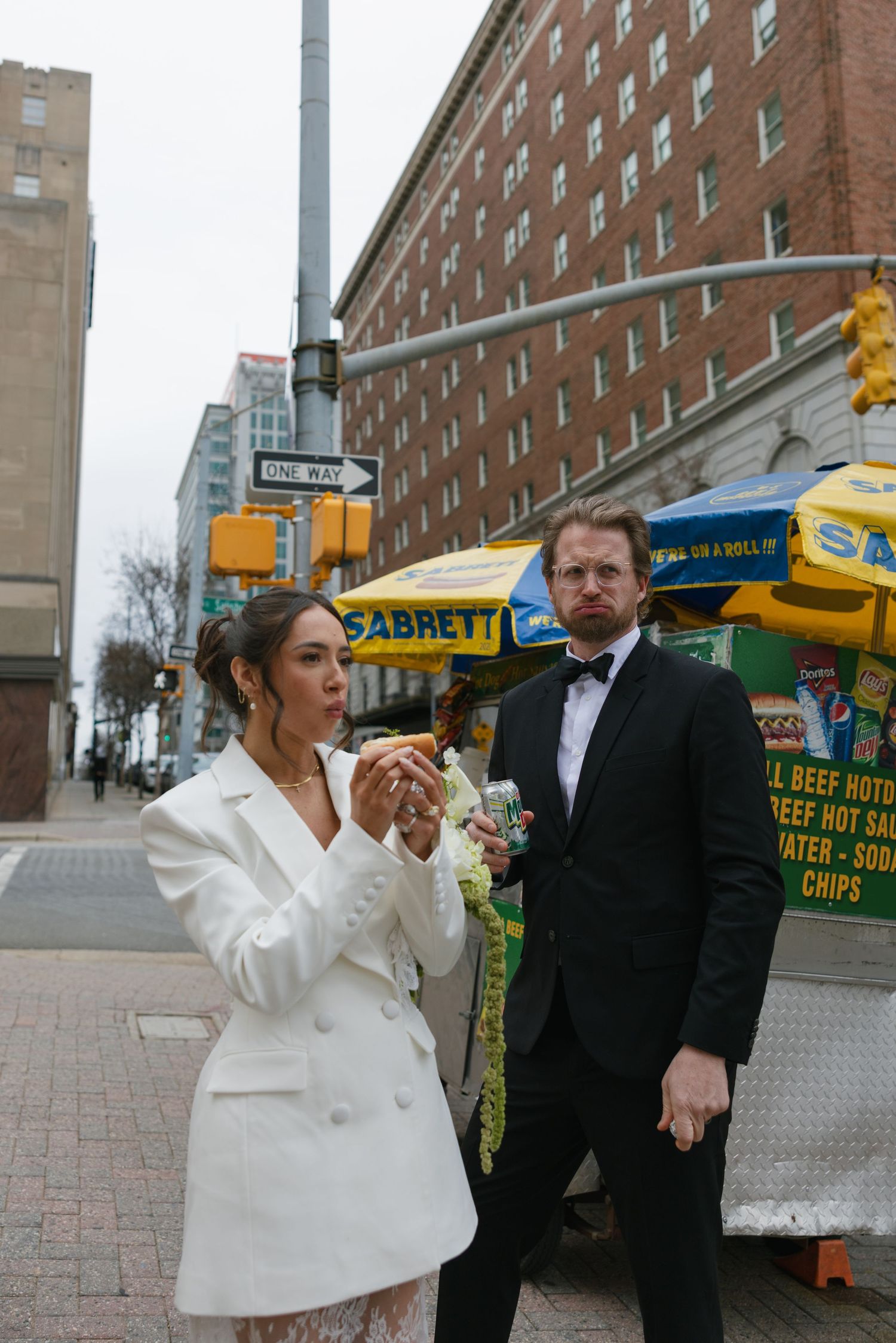 Bride in white blazer and groom in tuxedo eating hot dogs at a Sabrett street cart in New York City.