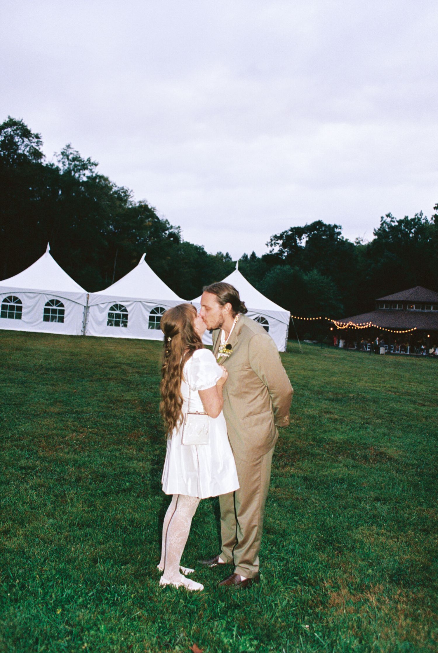 A couple shares a romantic moment on a grassy field with white event tents in the background.