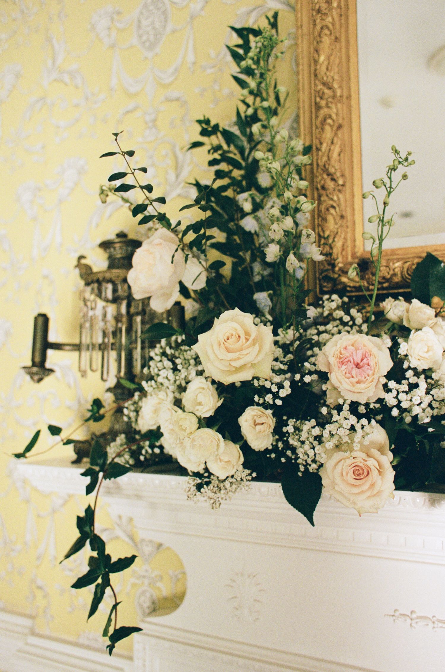 Cream and blush roses with baby's breath and greenery arranged on a white mantel beside a gilded mirror and yellow damask wallpaper.