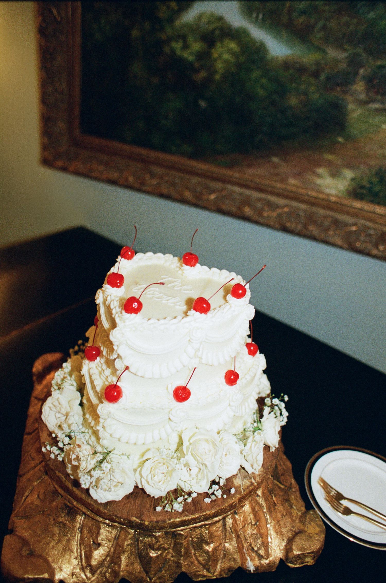 Two-tier white wedding cake with piped frosting, maraschino cherries and white roses on a gilded stand beside a plate and fork.