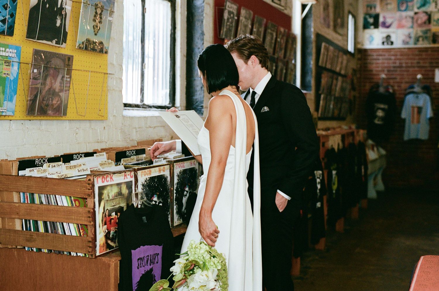 Bride in a white gown holding a bouquet kisses her groom in a dark suit while browsing vinyl records in a cozy record store.