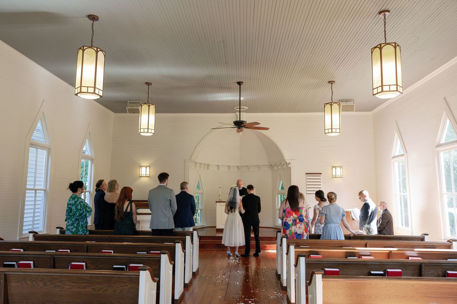 Wedding couple stands at the altar of a small white chapel with wooden pews as guests watch during a sunlit ceremony.