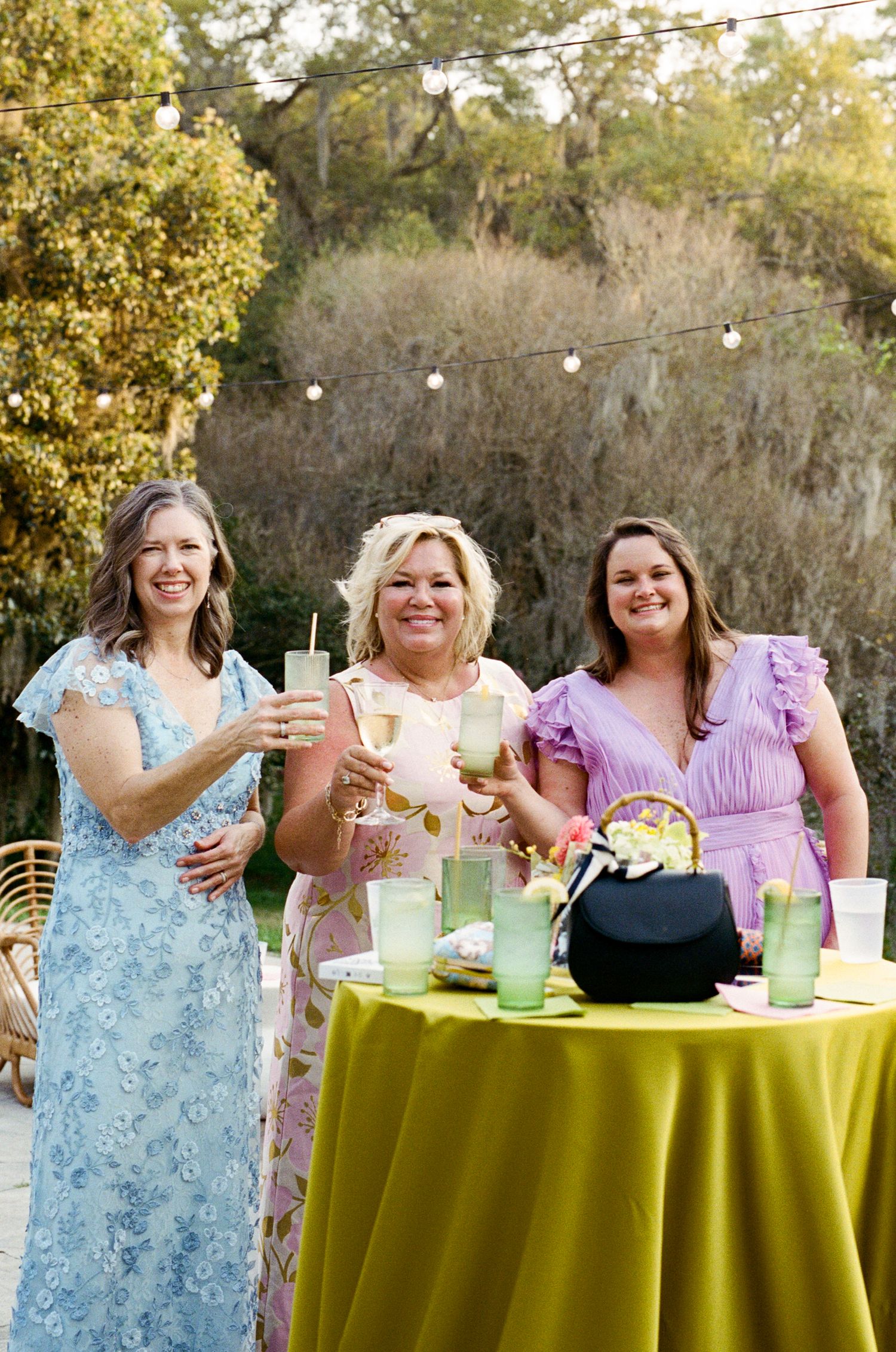 Three women in colorful dresses toasting drinks at an outdoor garden party with string lights and a green tablecloth.
