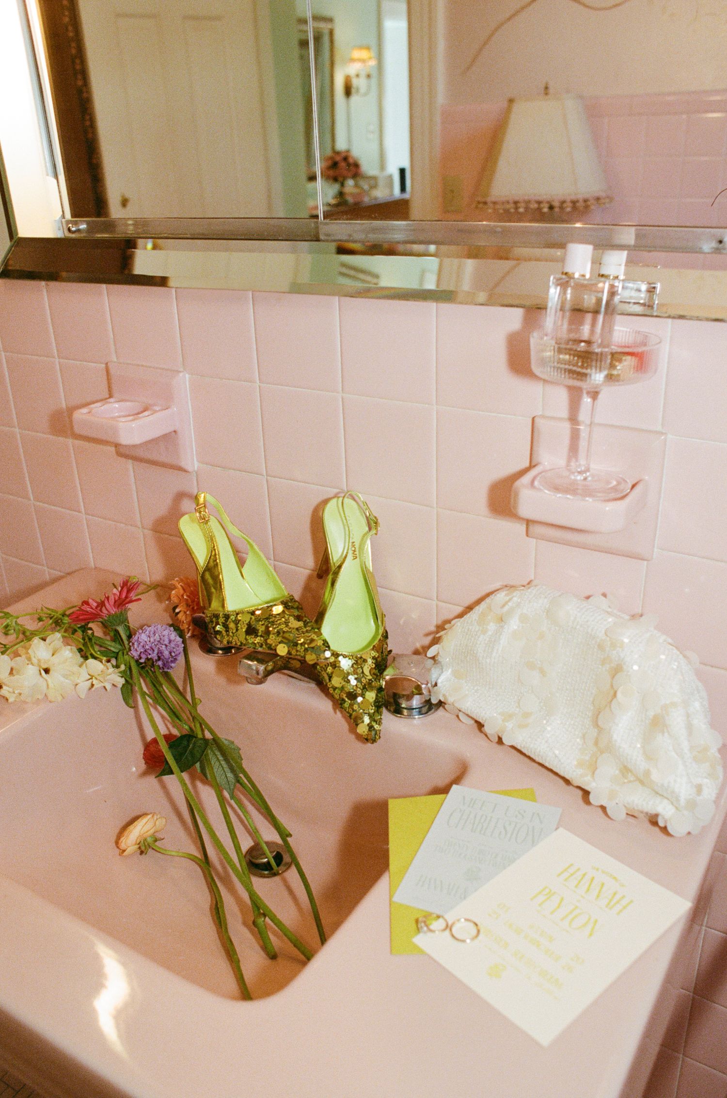 Pink vintage bathroom sink styled with green heels, flowers, a wine glass, and wedding invitation for a bridal shoot.
