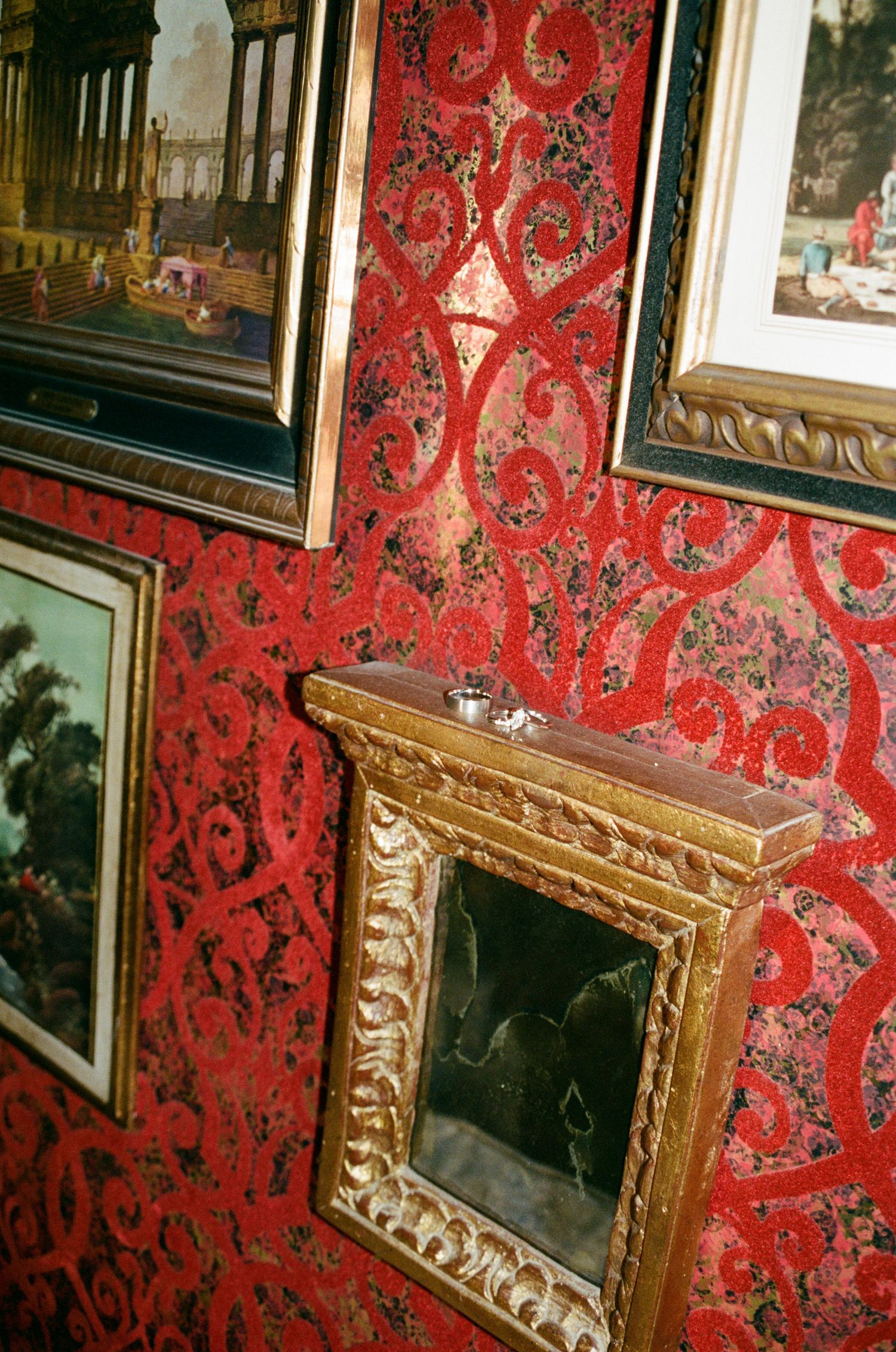 Ornate gold-framed artwork hangs on vibrant red damask wallpaper in an elegant gallery setting.
