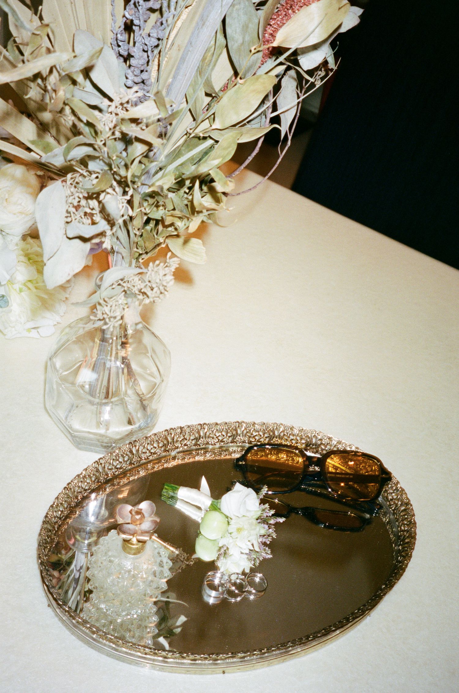 Ornate silver mirrored tray with decorative edges reflecting a glass vase with delicate white flowers on a light surface.
