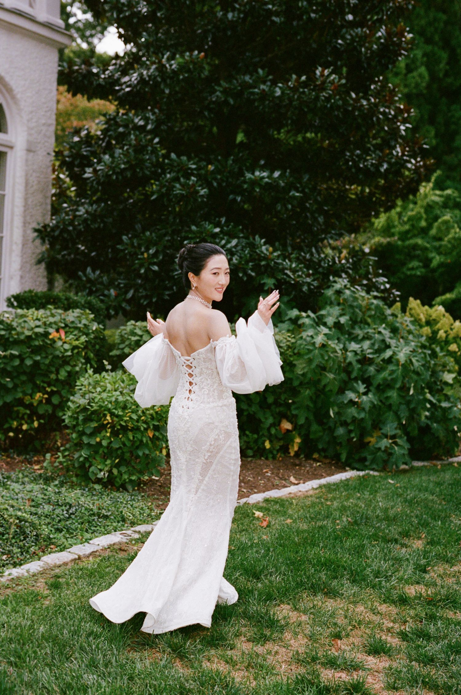 A bride in a white lace wedding gown with billowing sleeves poses gracefully in a garden setting.