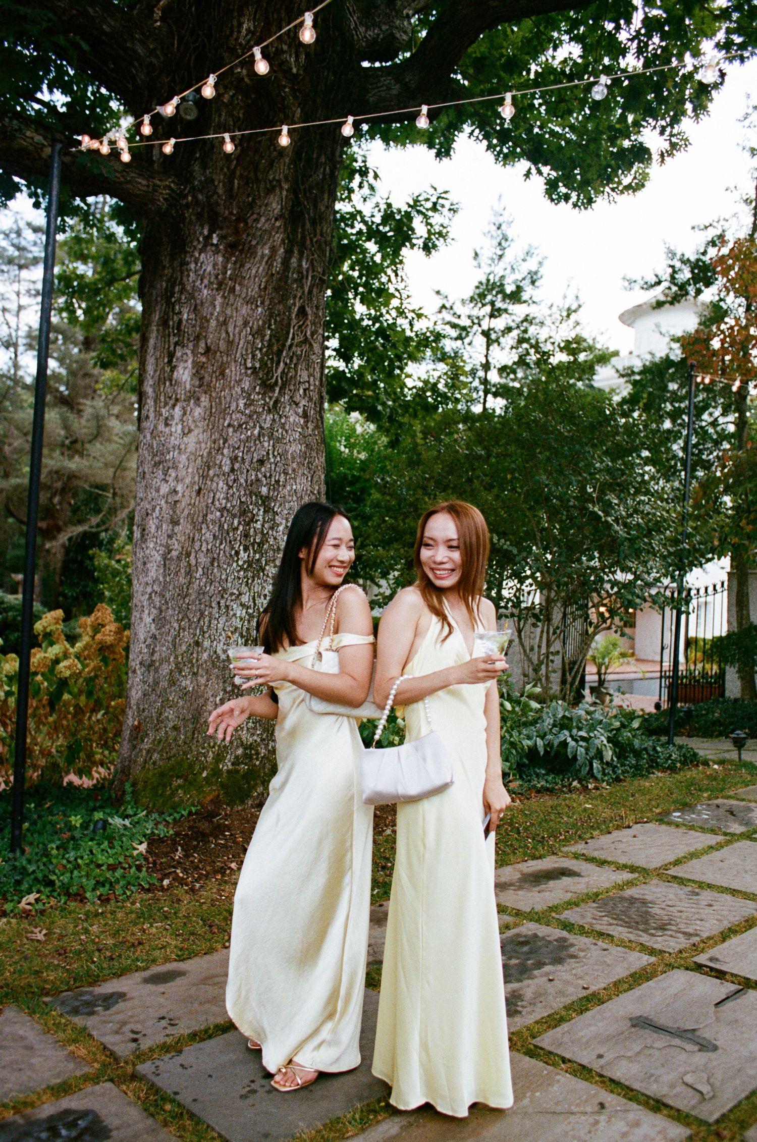 Two people in cream-colored dresses laugh and dance together on a stone patio under string lights and trees.