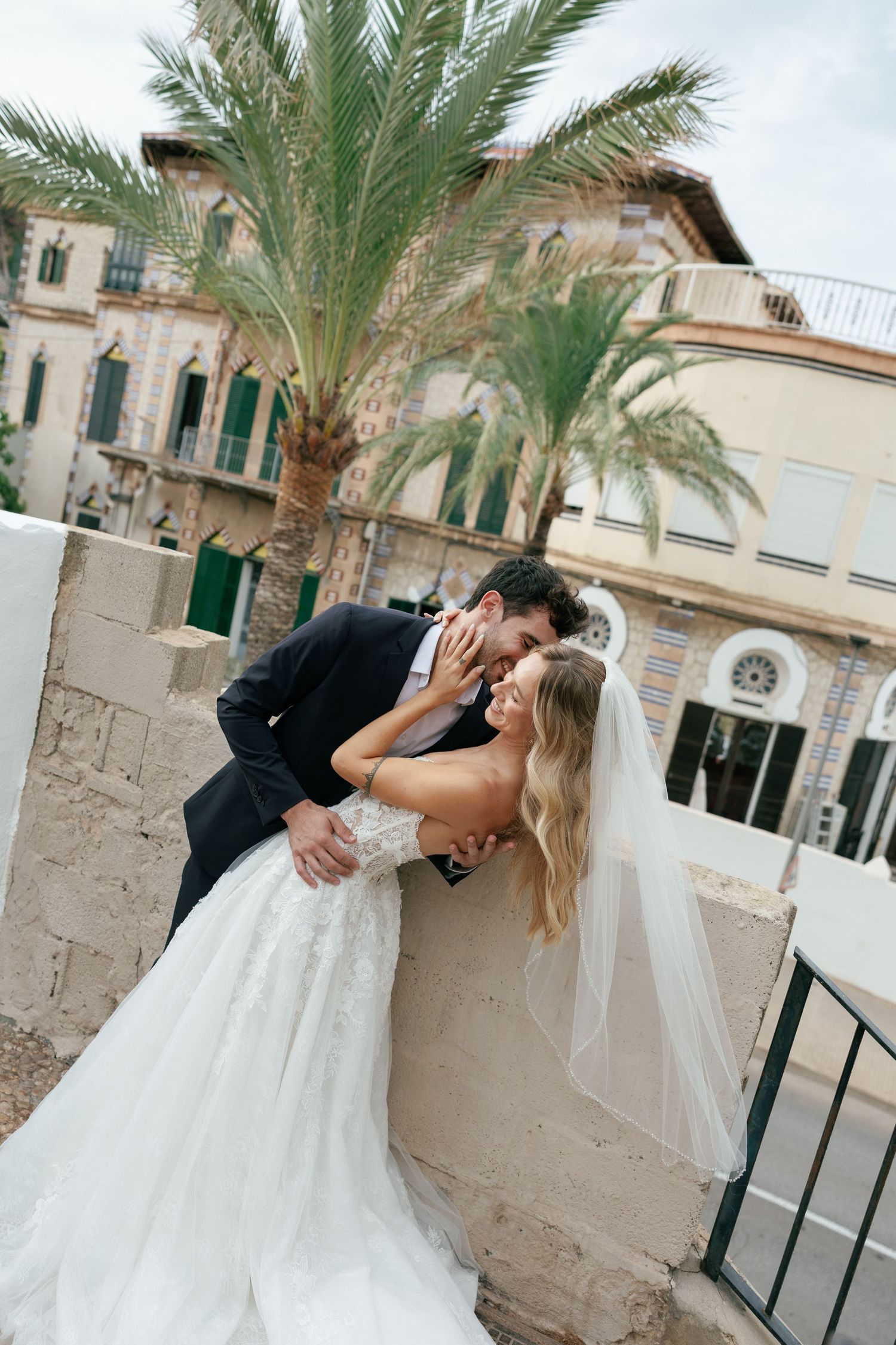 A romantic wedding couple dips and kisses beneath palm trees in a Mediterranean courtyard setting.