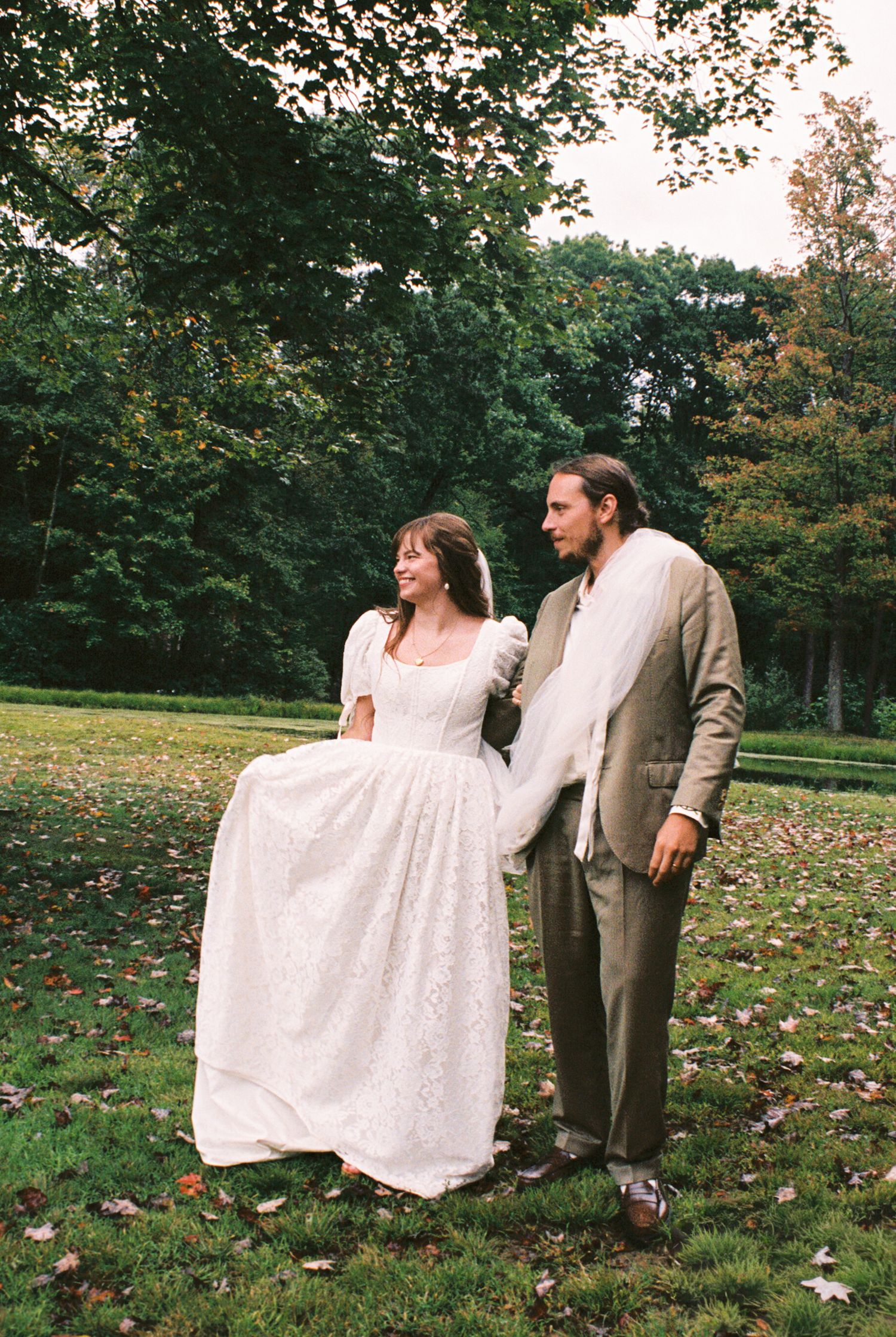 A couple in vintage-style wedding attire stands together on an autumn lawn surrounded by trees and fallen leaves.