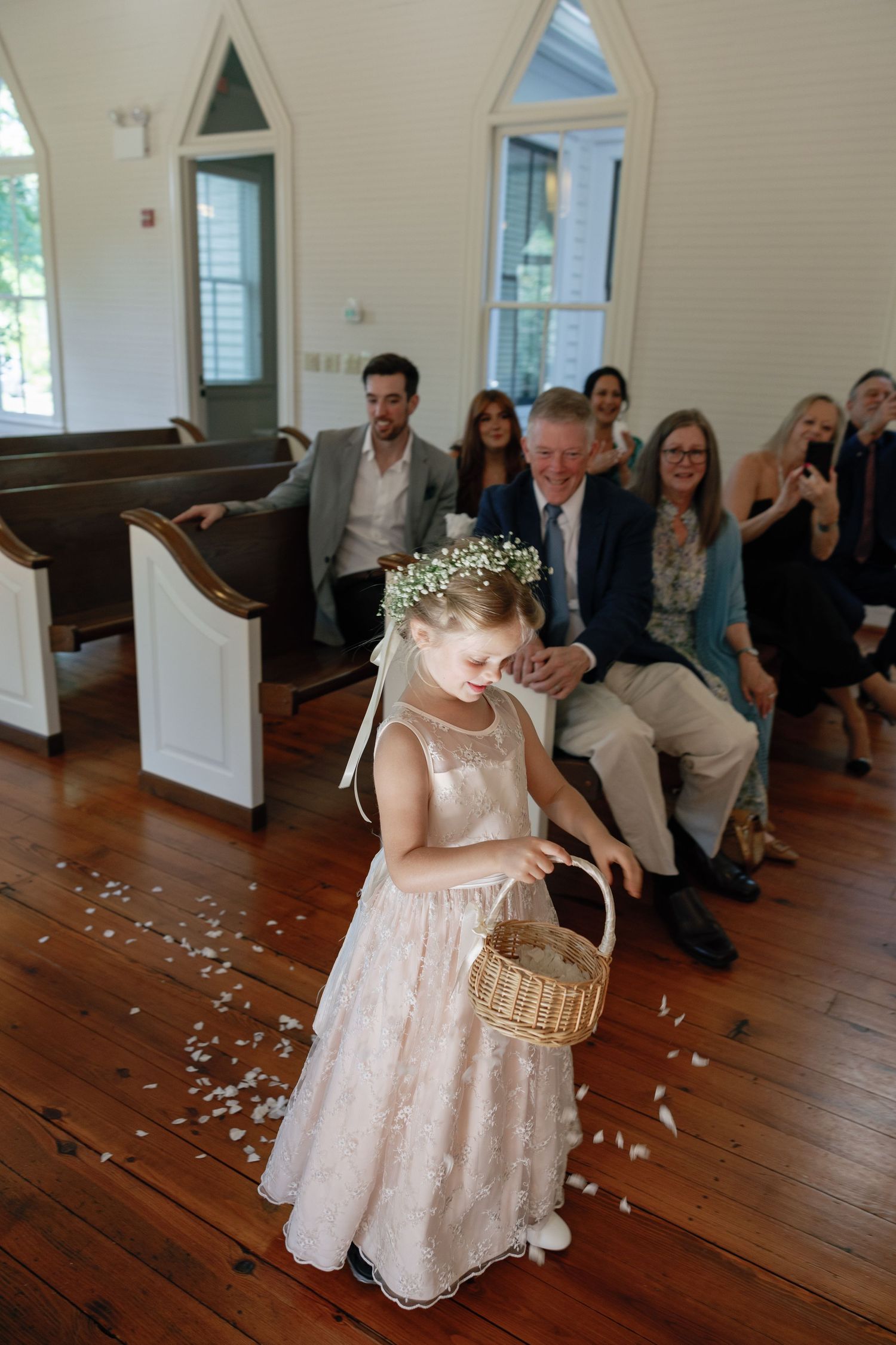 Flower girl in a blush dress and floral crown carries a wicker basket, scattering petals down the chapel aisle as wedding guests smile.