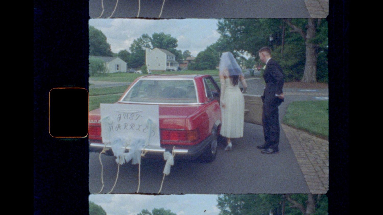 Bride in a white dress and veil stands beside a red vintage car with a just married sign as a groom in a dark suit waits nearby.
