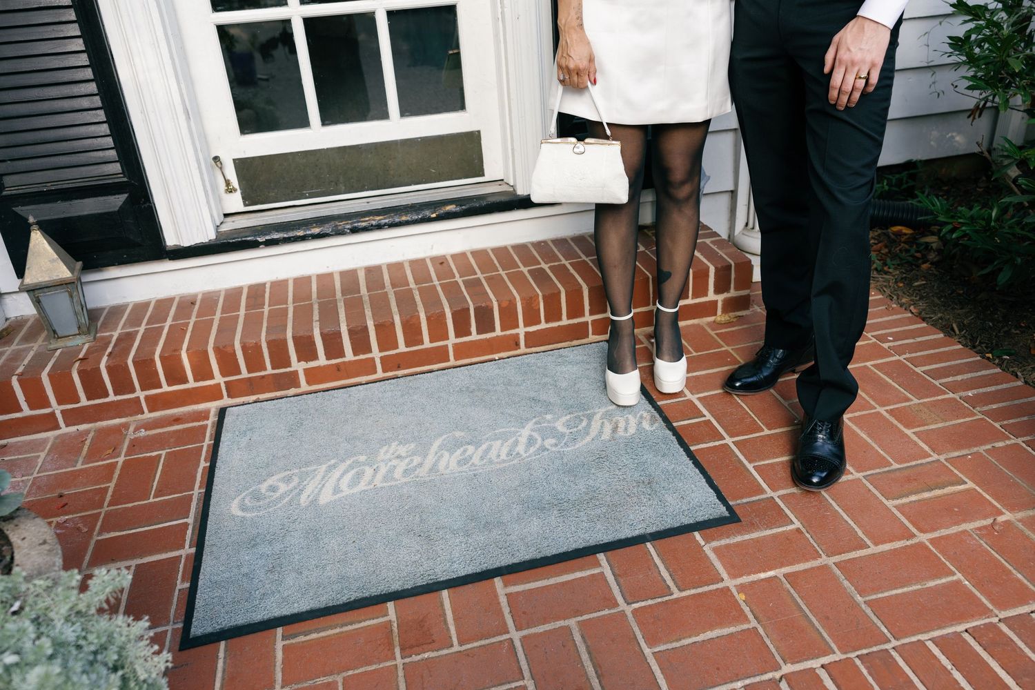 Couple standing on a brick porch at a white door, with a gray welcome mat reading Matchbook Inn at their feet.